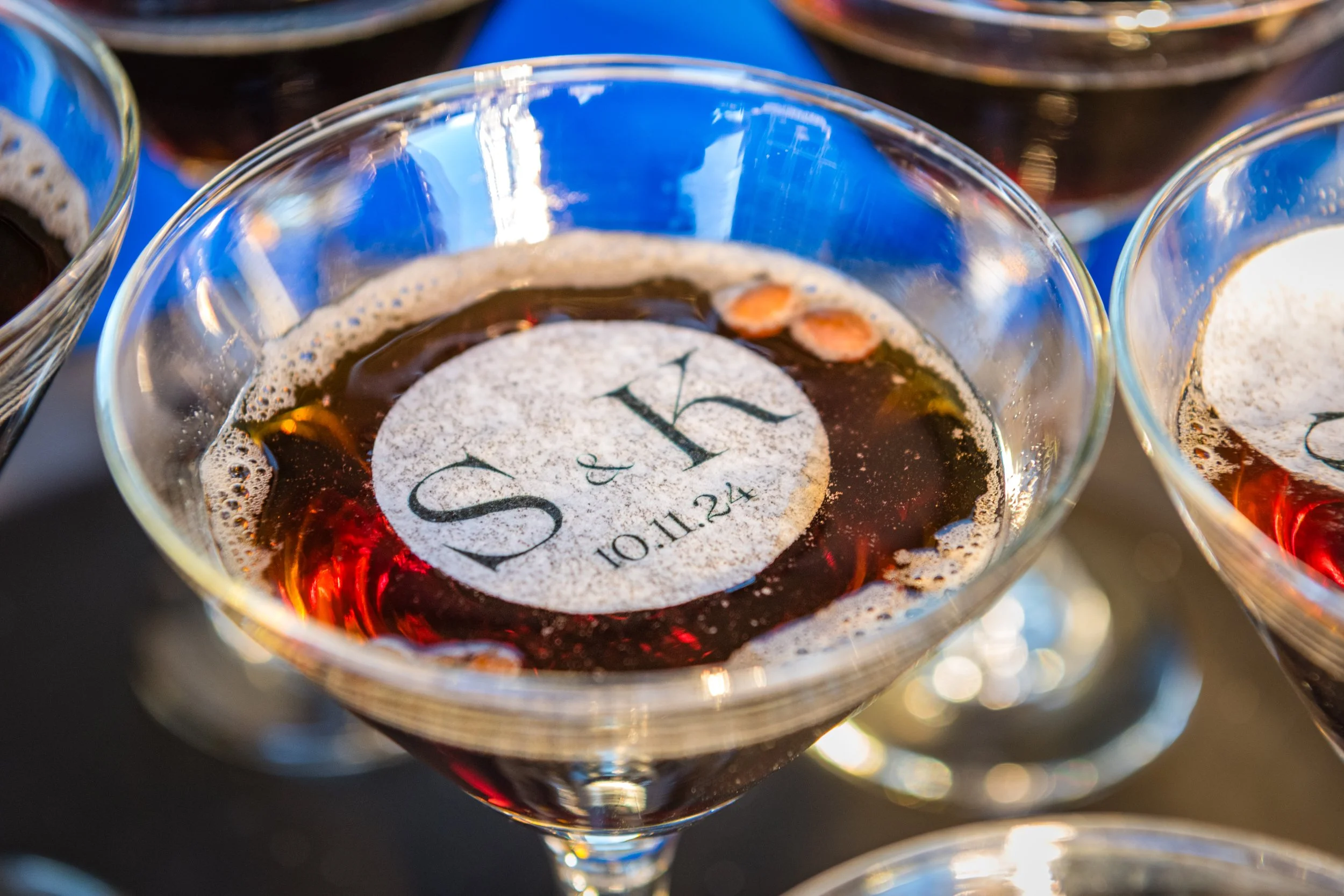 Close-up of a glass of red wine with a white round sticker on the surface that has the word 'S' and a date '10.11.24' written on it, with blurred glasses in the background.