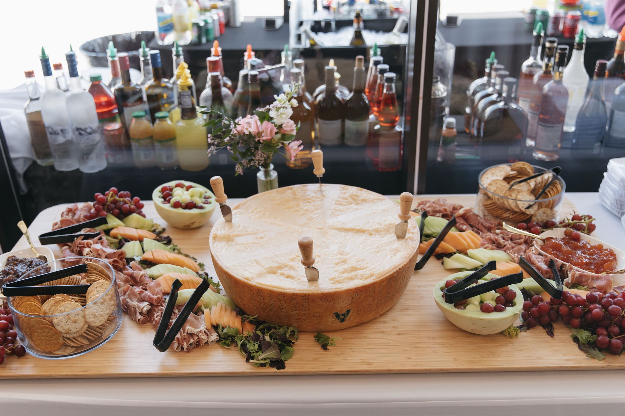 A table with assorted cheeses, crackers, grapes, sliced meats, and a wheel of cheese with knives, in front of a display of various bottled sauces and condiments.