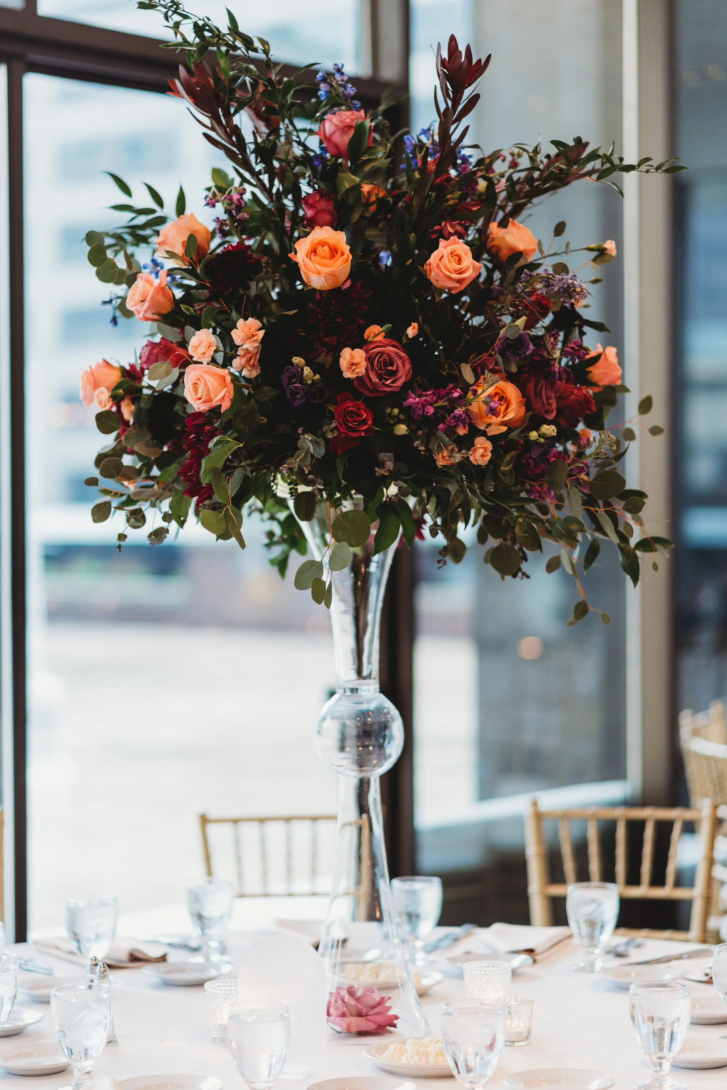 A tall glass vase holding a large arrangement of mixed flowers including roses, greenery, and other blooms, set on a white tablecloth with glassware and table settings, near a window in a bright room.