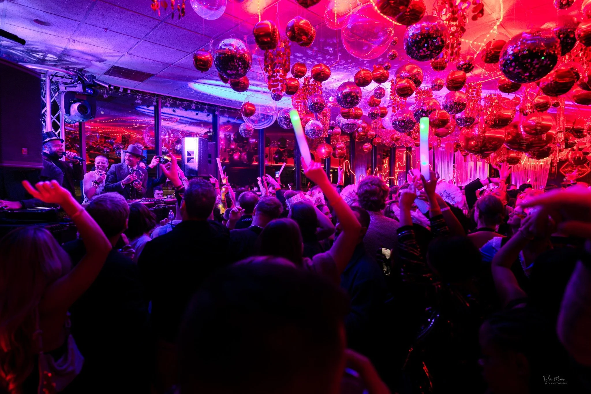 A lively party scene with colorful disco balls hanging from the ceiling and a crowd of people dancing. There are vibrant DJ lights and decorations creating a festive atmosphere.
