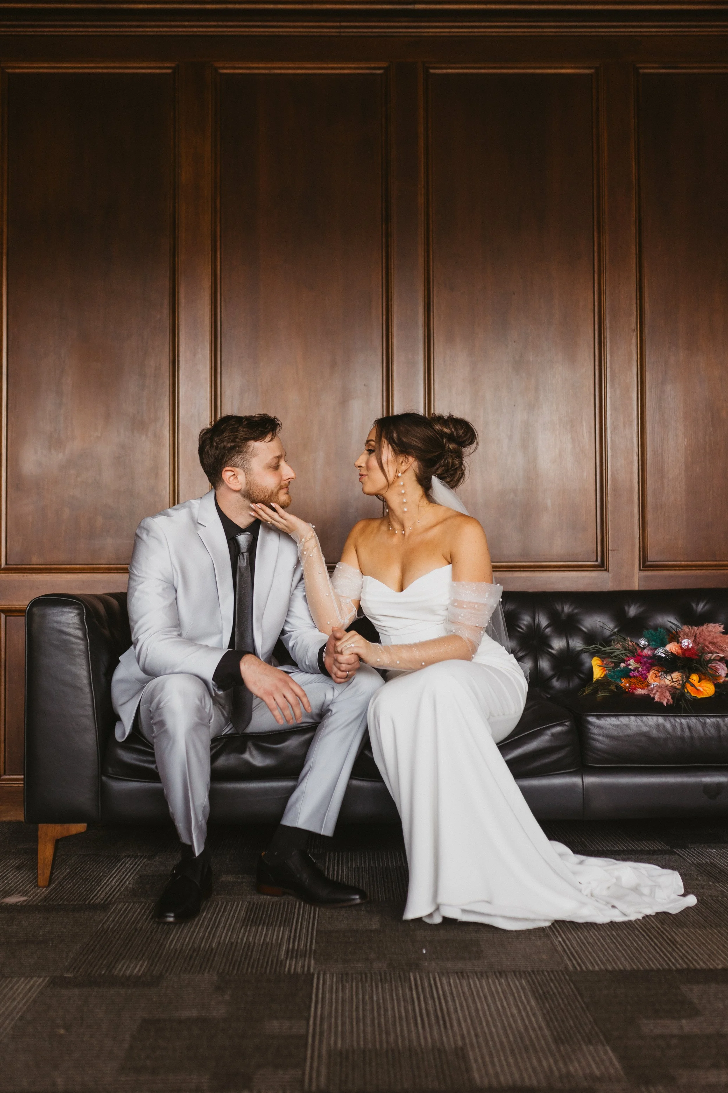 Newlyweds on a dark leather sofa in the executive bridal suite at JPS Events at Regions Tower. Features dark wood paneling, private prep suites, zero late-out fees, & built-in owner day-of coordination.