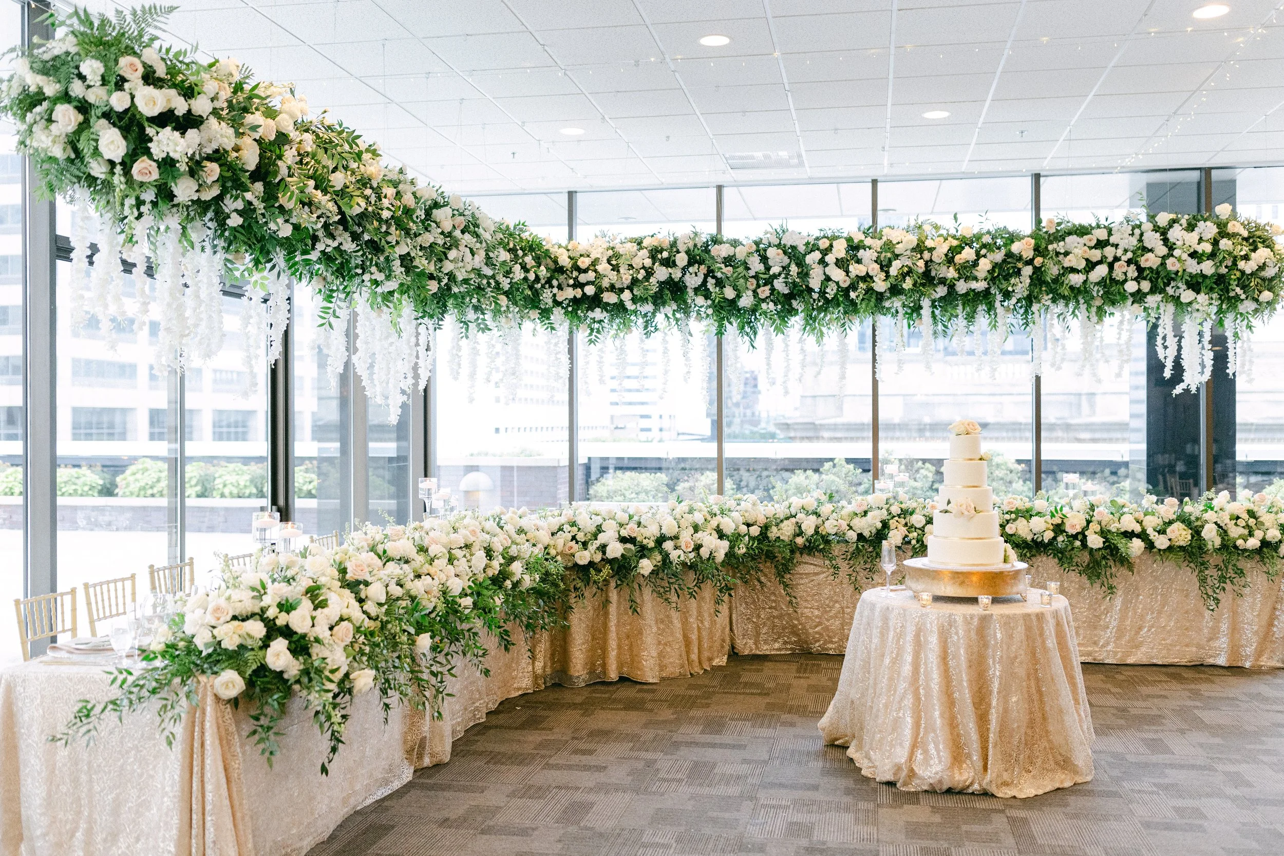 An elegant wedding reception setup with a long floral arch of white and blush roses, greenery, and cascading white flowers, set in a glass-walled venue with city skyline views. A tiered white wedding cake is displayed on a round table covered in a lace tablecloth, surrounded by floral arrangements and glassware.