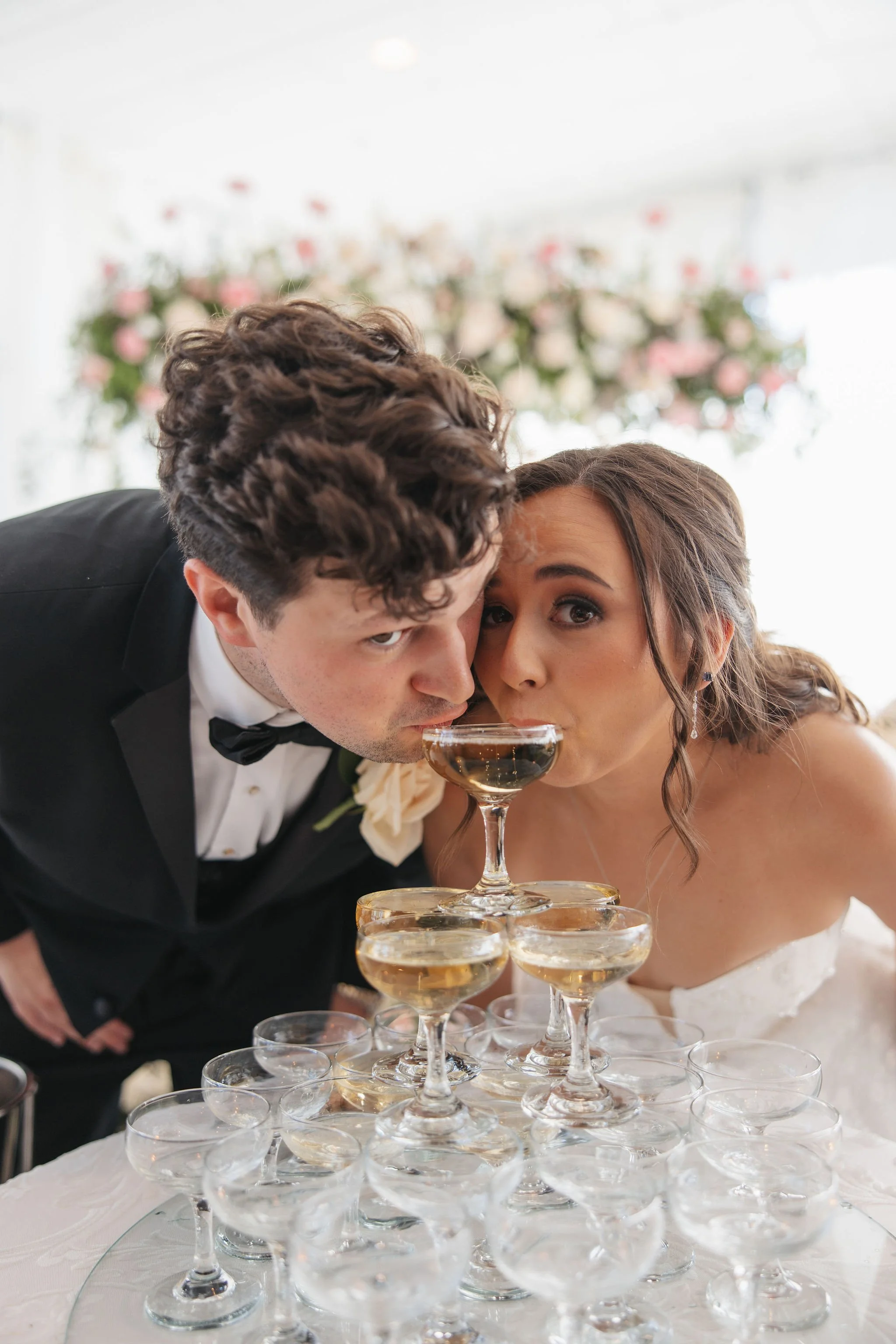A newlywed couple with a man in a tuxedo and a woman in a wedding dress, leaning over a pyramid of champagne glasses, at their wedding reception with a floral backdrop.