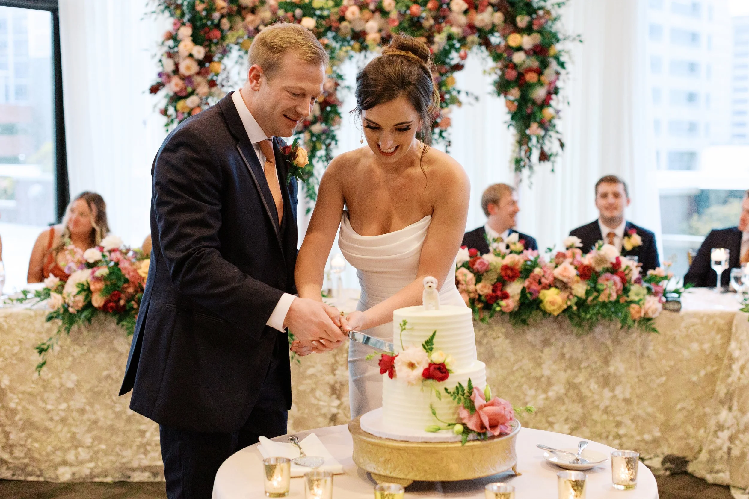A bride and groom cutting a wedding cake together at their reception, with guests and floral decorations in the background.