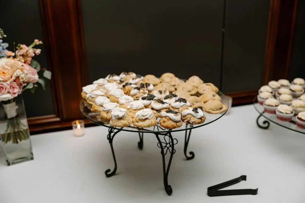 Assorted cookies on a glass platter, displayed on a black wrought iron stand at a dessert table, with cupcakes in the background and a flower arrangement on the side.