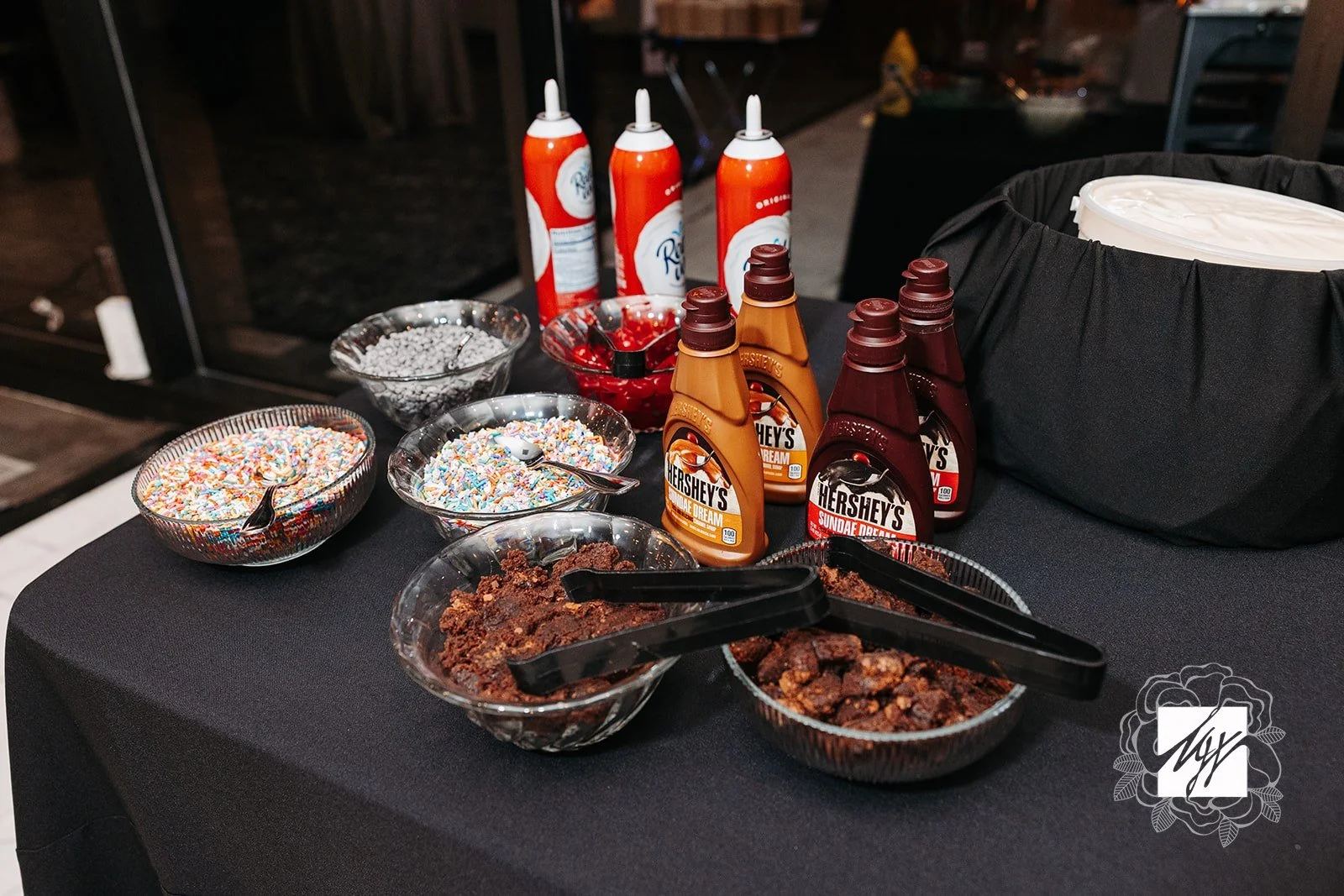 A table with various toppings and syrups for ice cream, including sprinkles, chocolate chunks, Hershey's chocolate syrup, caramel syrup, and whipped cream—late-night snacks by luxury in-house catering at JPS Events at Regions Tower in Indy.  
