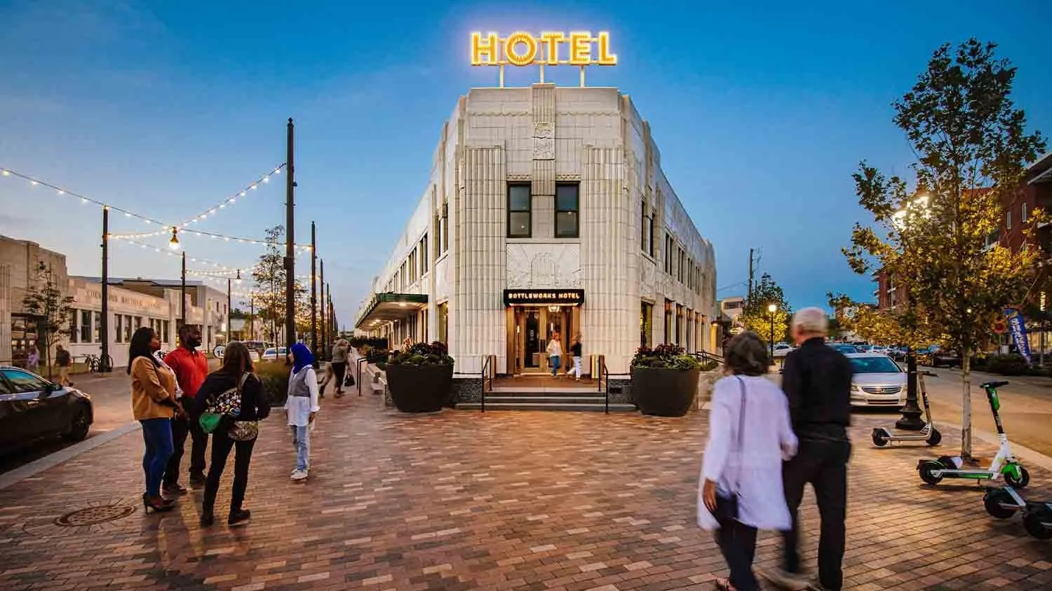 A street scene at dusk featuring a corner hotel with a neon sign reading 'HOTEL'. People are walking and standing on the brick sidewalk, with some electric scooters parked nearby. The hotel building has a classic facade with an art deco style, and string lights are hanging across the street.