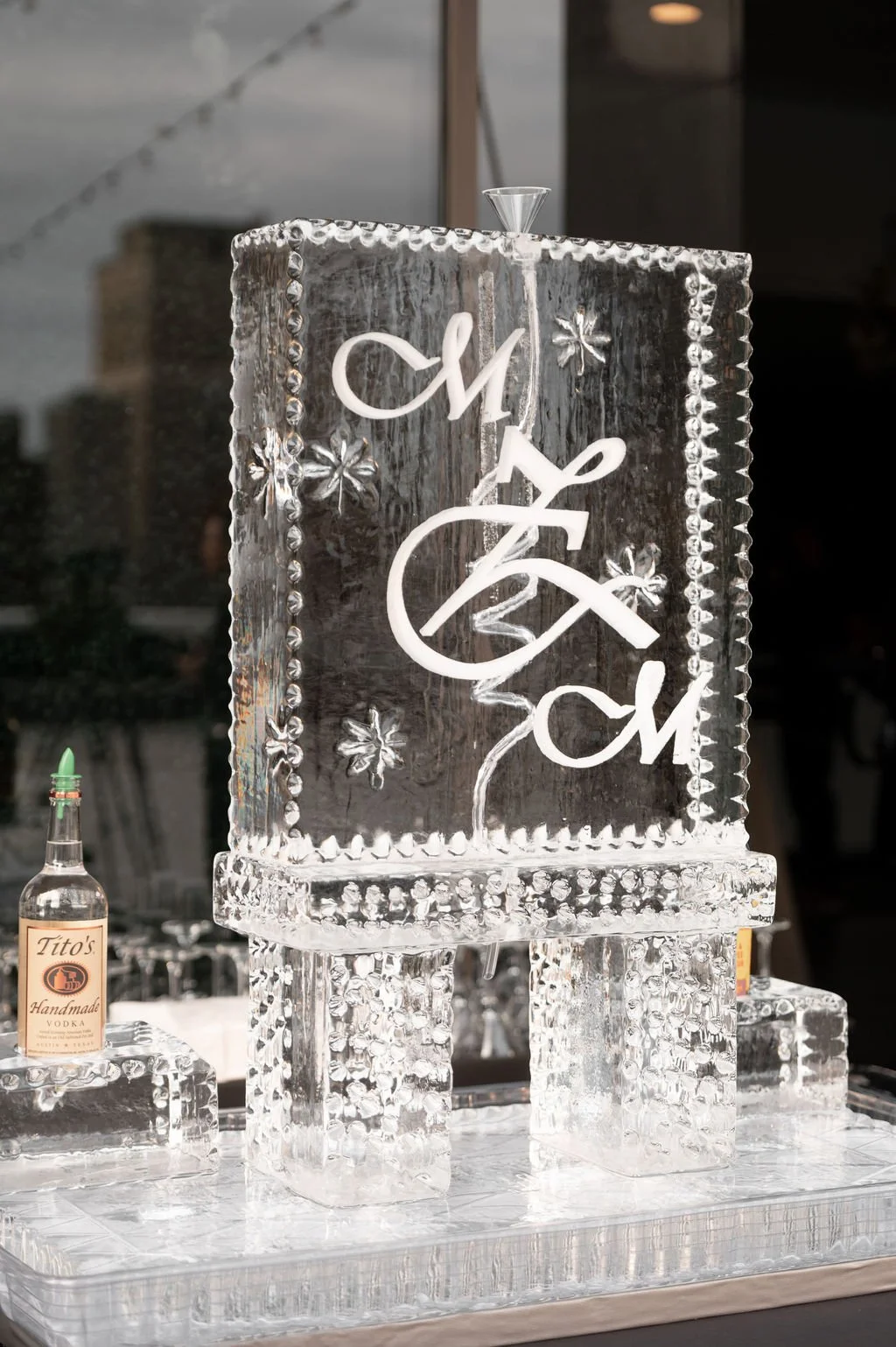 A decorative ice sculpture with the words 'N + Z M' engraved, along with floral patterns, placed on a table. A small bottle of Tito's Handmade Vodka is positioned near the sculpture.