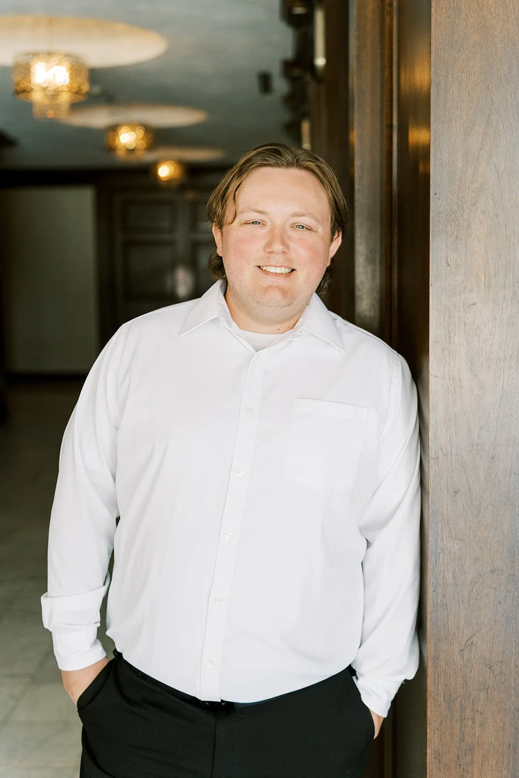 A man with medium-length brown hair wearing a white dress shirt standing indoors, leaning against a wooden wall, smiling at the camera.