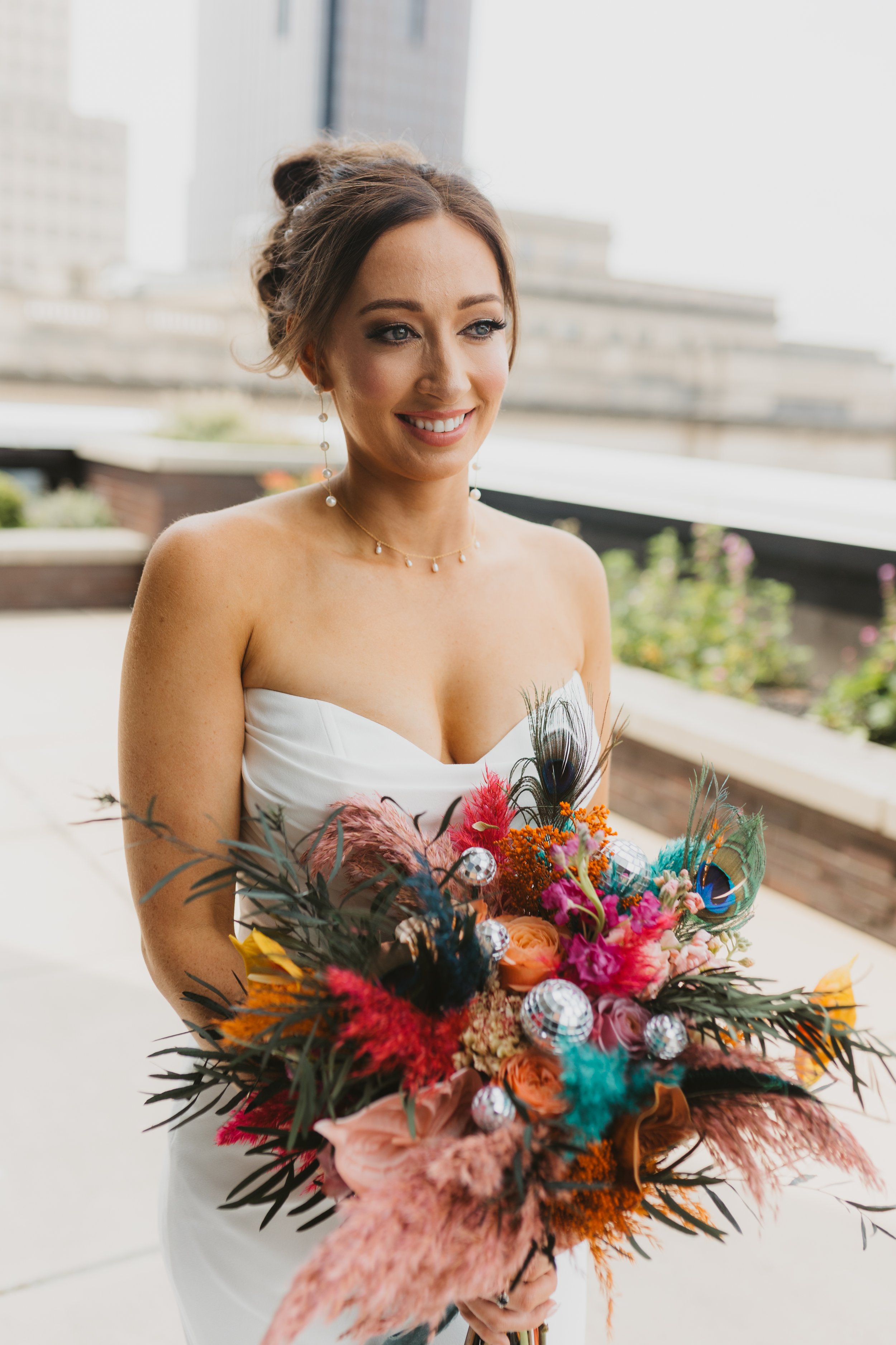 A woman in a strapless white dress holding a colorful bouquet of flowers and peacock feathers, smiling, with a cityscape in the background.