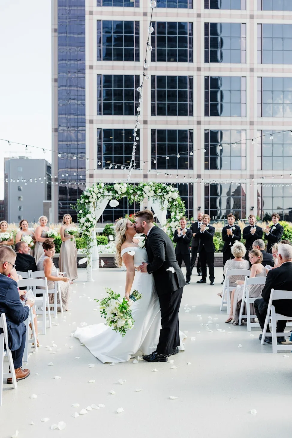 A wedding ceremony taking place outdoors on a rooftop with a modern glass building in the background. The bride and groom are kissing, surrounded by guests, floral arrangements, and string lights.