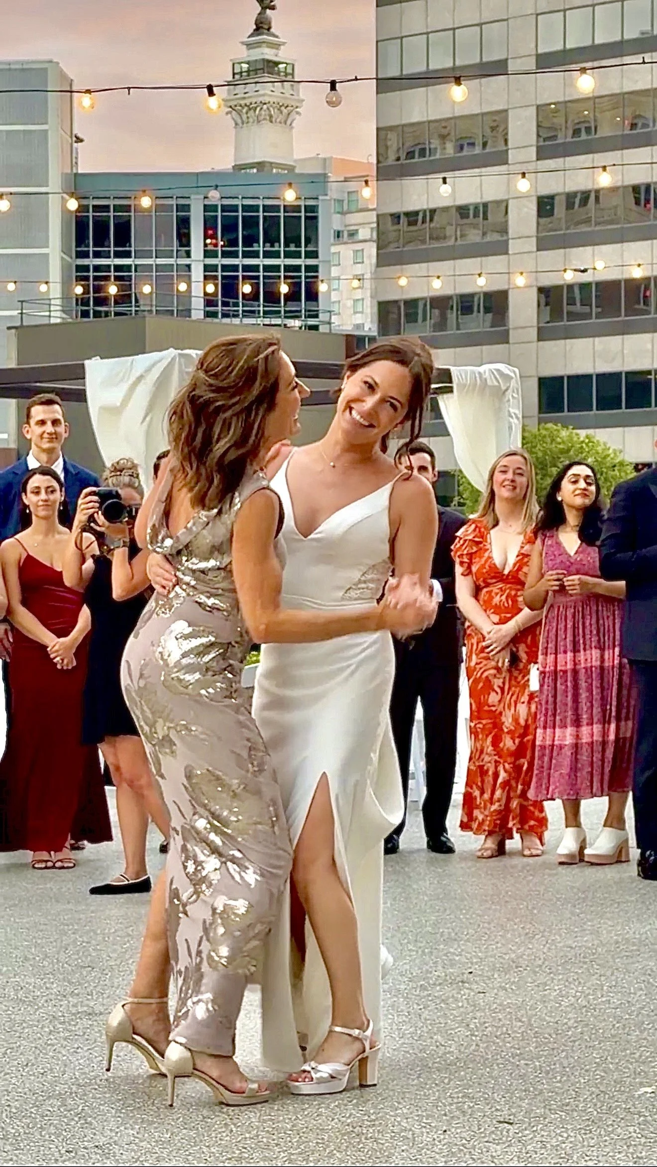 A bride and her mom dance at an outdoor wedding celebration on the private rooftop terrace at JPS Events at Regions Tower in Downtown Indianapolis, with string lights overhead and illuminated city buildings surrounding the dance floor.