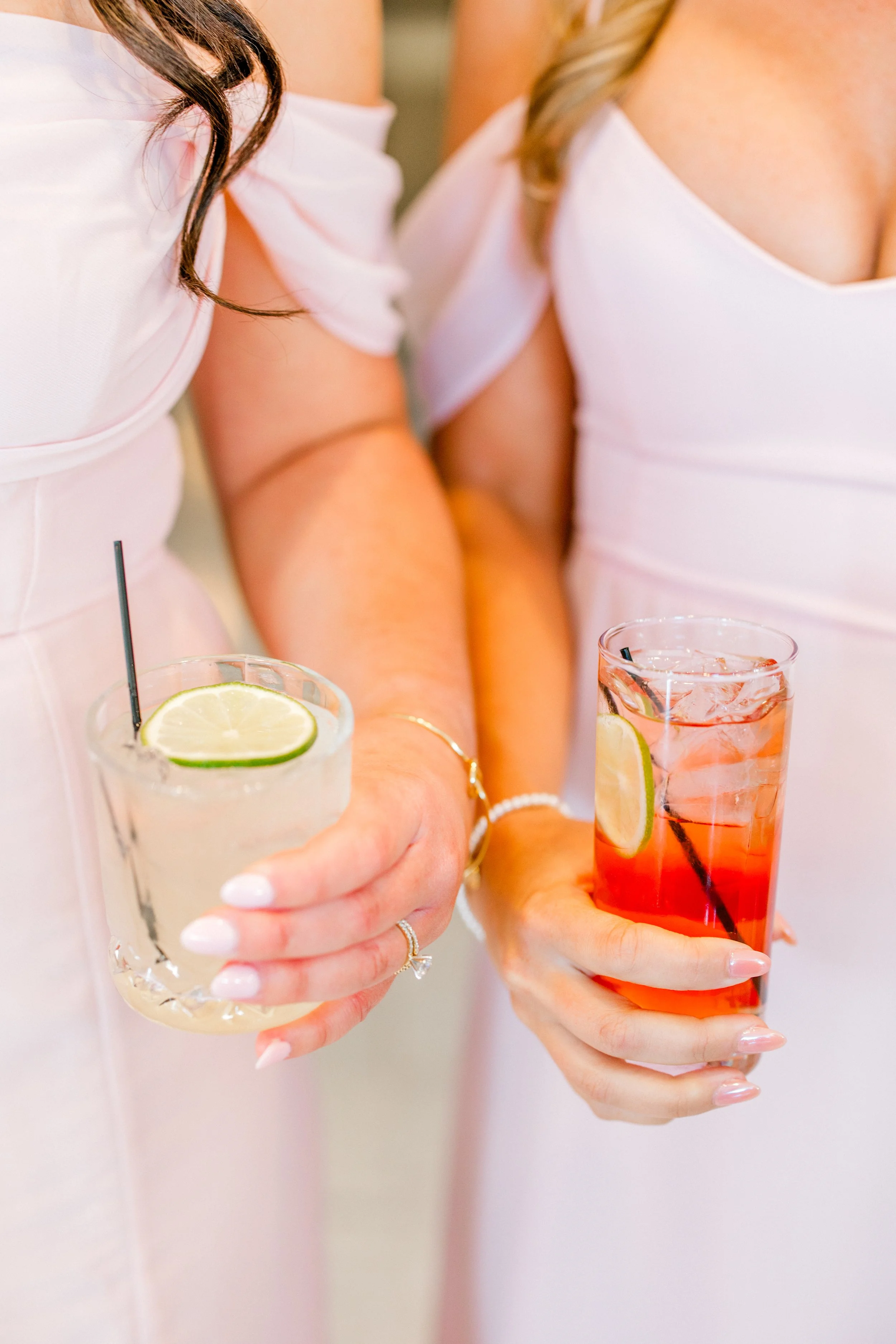 Two women in white dresses holding colorful drinks with lime slices, ice, and straws.