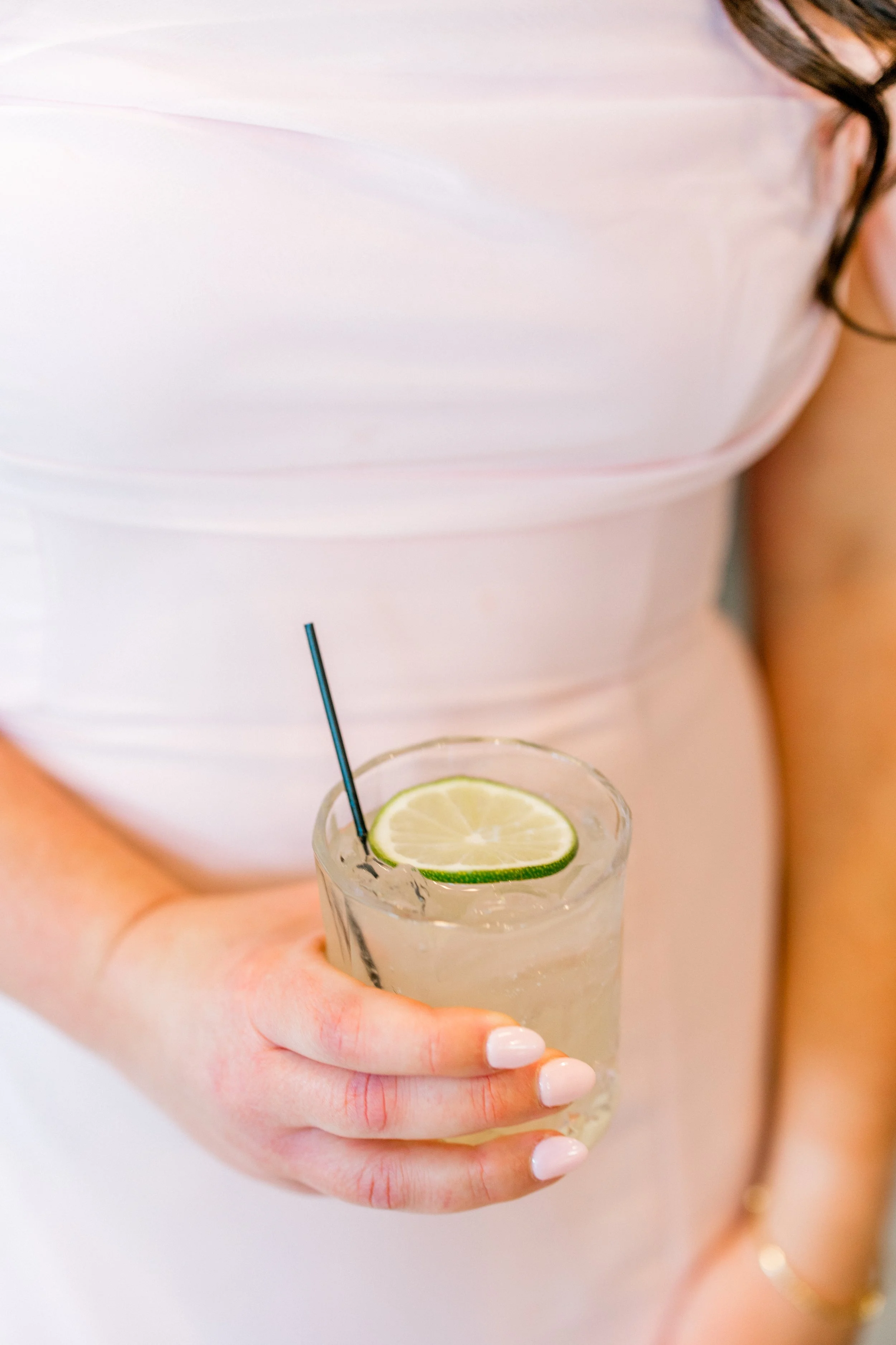 Person holding a glass of clear beverage with lime slices, a black straw, and a white top