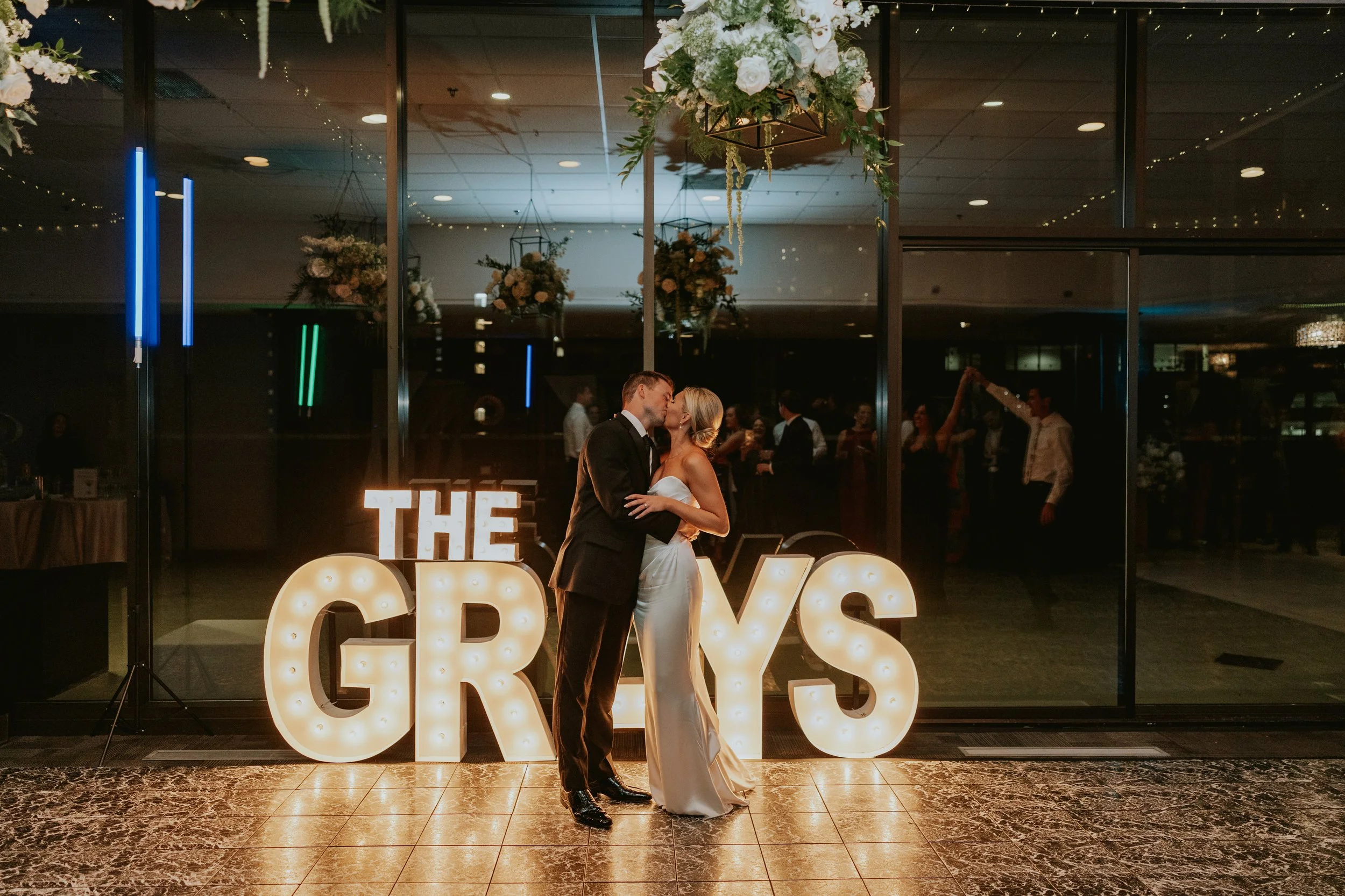 A newlywed couple sharing a kiss during their wedding reception, with illuminated marquee letters spelling 'THE GRAYS' in the background.