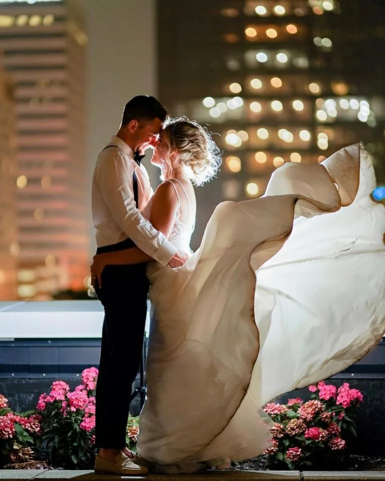 A couple dressed in wedding attire standing close together at night in an urban setting, with city lights in the background. The bride is holding her dress out while they share an intimate moment.