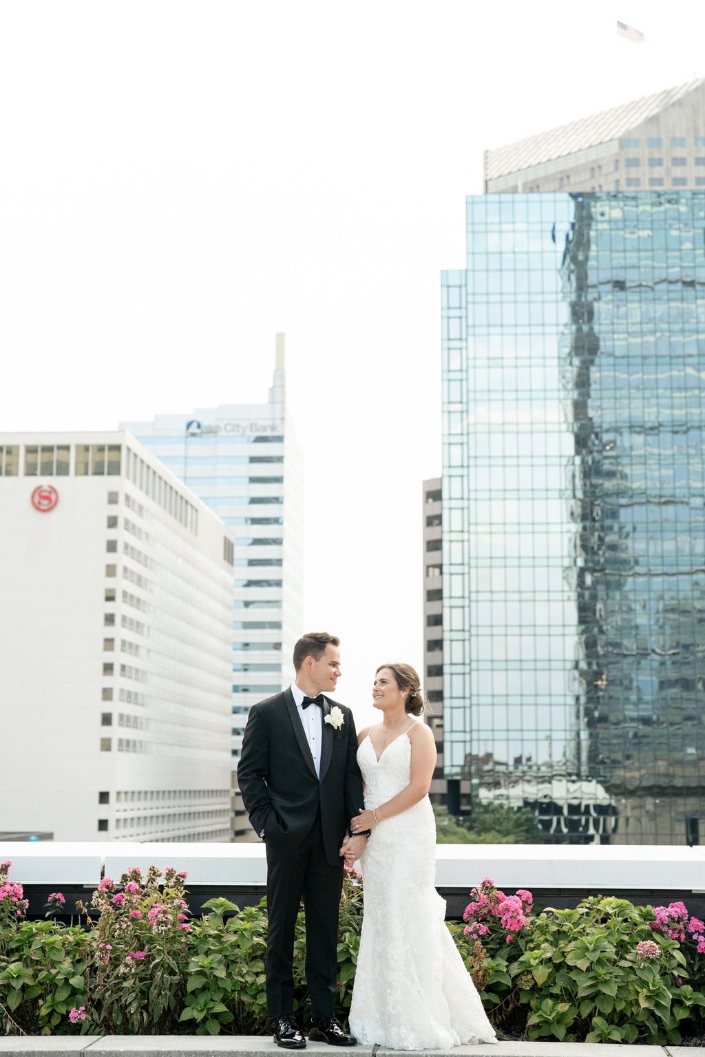 A newlywed couple smiling on the private rooftop garden terrace at JPS Events at Regions Tower in Downtown Indianapolis, featuring custom in-house pink flowers and panoramic city skyline views in daylight.