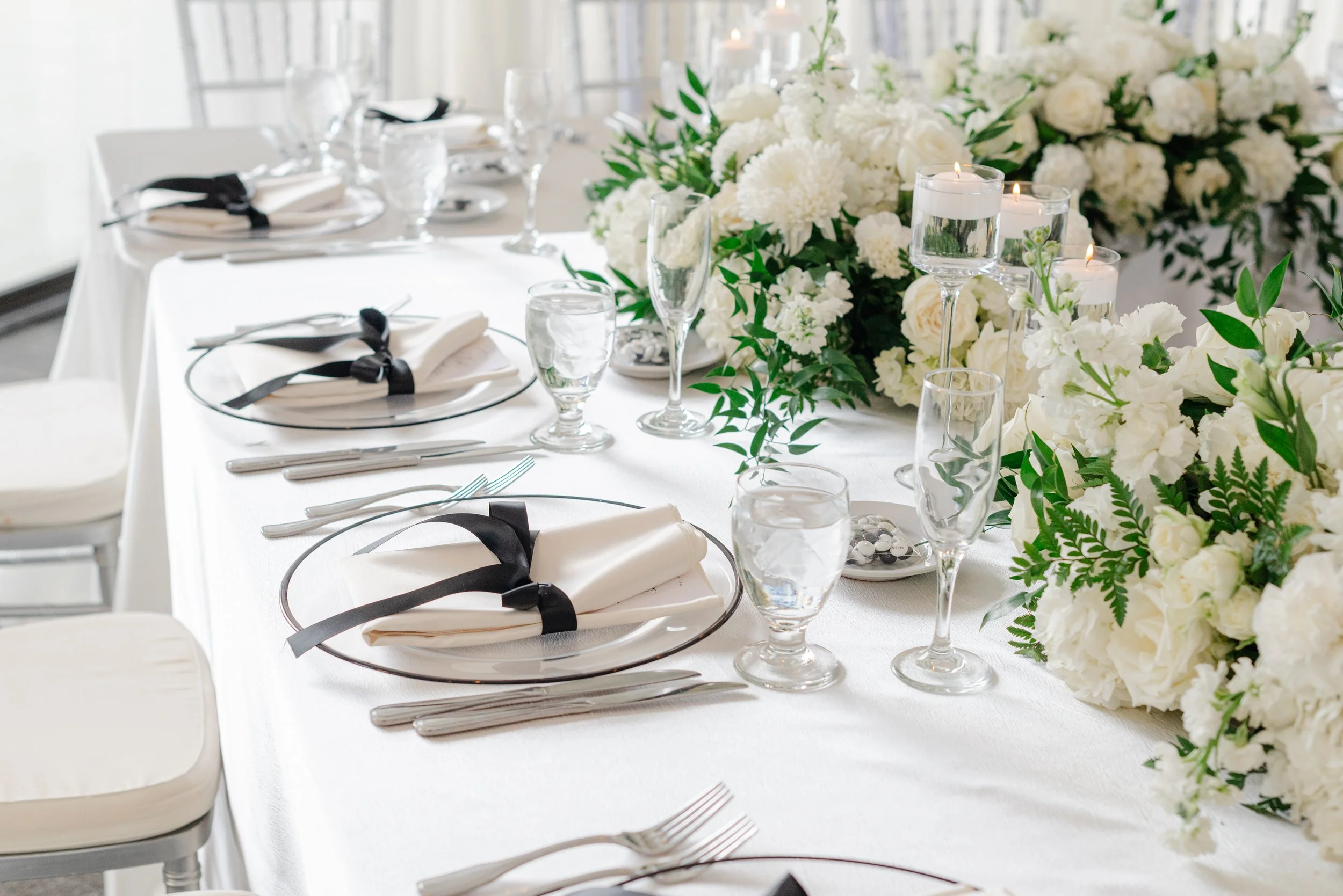 Elegant wedding table setting with white flowers, candles in glass holders, white napkins tied with black ribbons, and glassware on a white tablecloth.