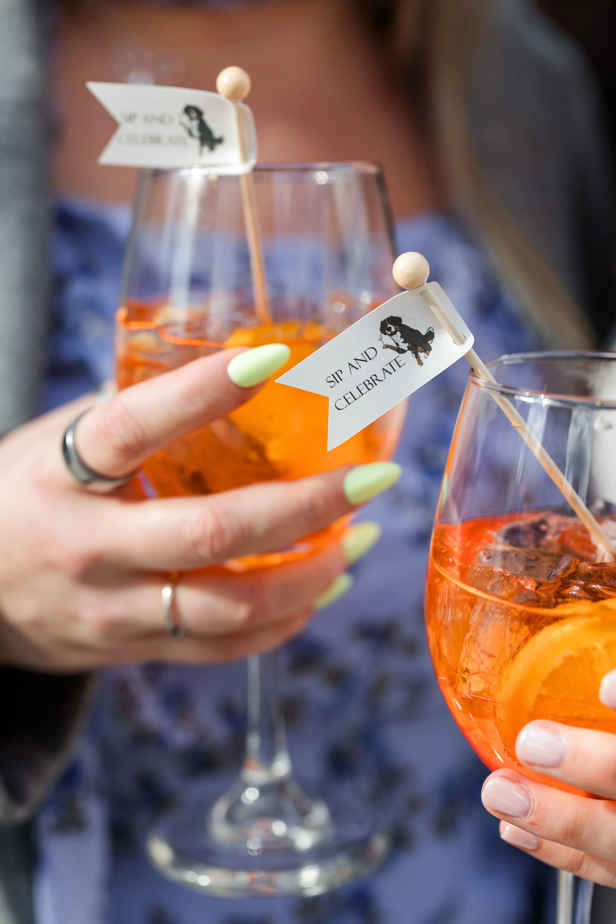 Close-up of a person holding an orange-colored drink in a wine glass, with a small flag that reads 'Sip and Celebrate' attached to a toothpick, with another glass in the background.