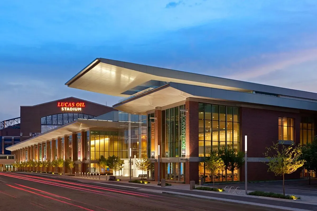 Modern stadium and convention center with large glass windows, trees, streetlights, and an illuminated Lucas Oil Stadium sign in the background at dusk.