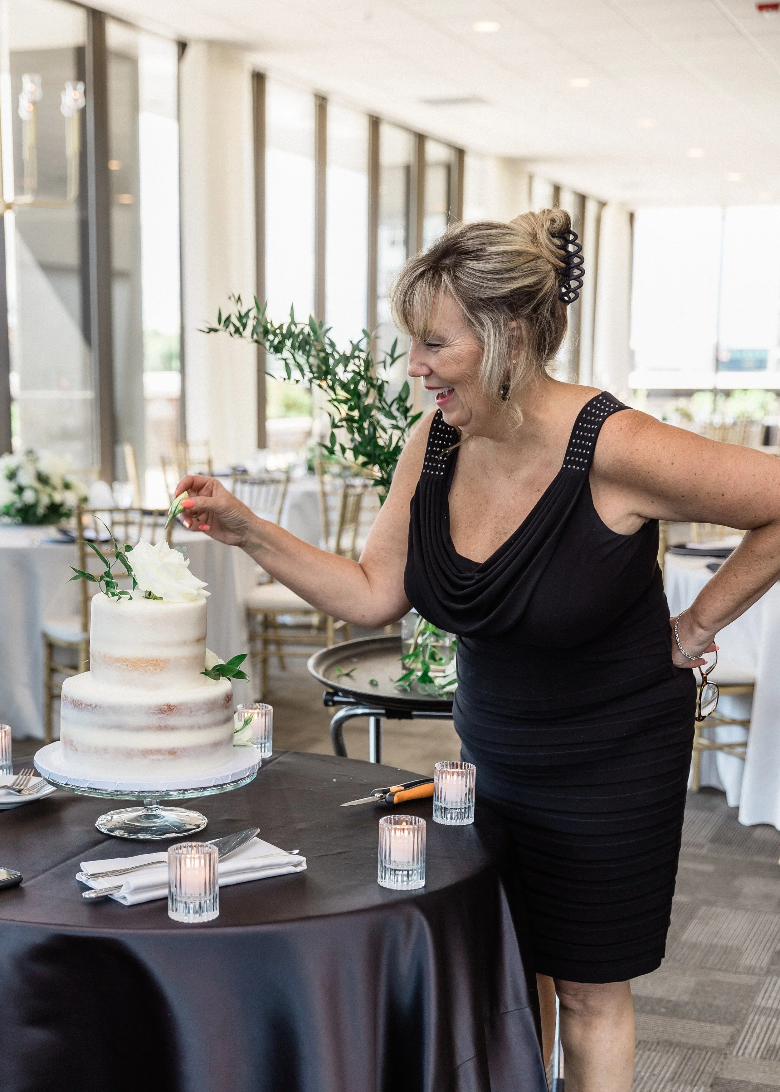 A woman in a black dress decorating a white, three-tiered wedding cake with flowers. She is smiling and leaning over the cake table in a bright, modern event space.