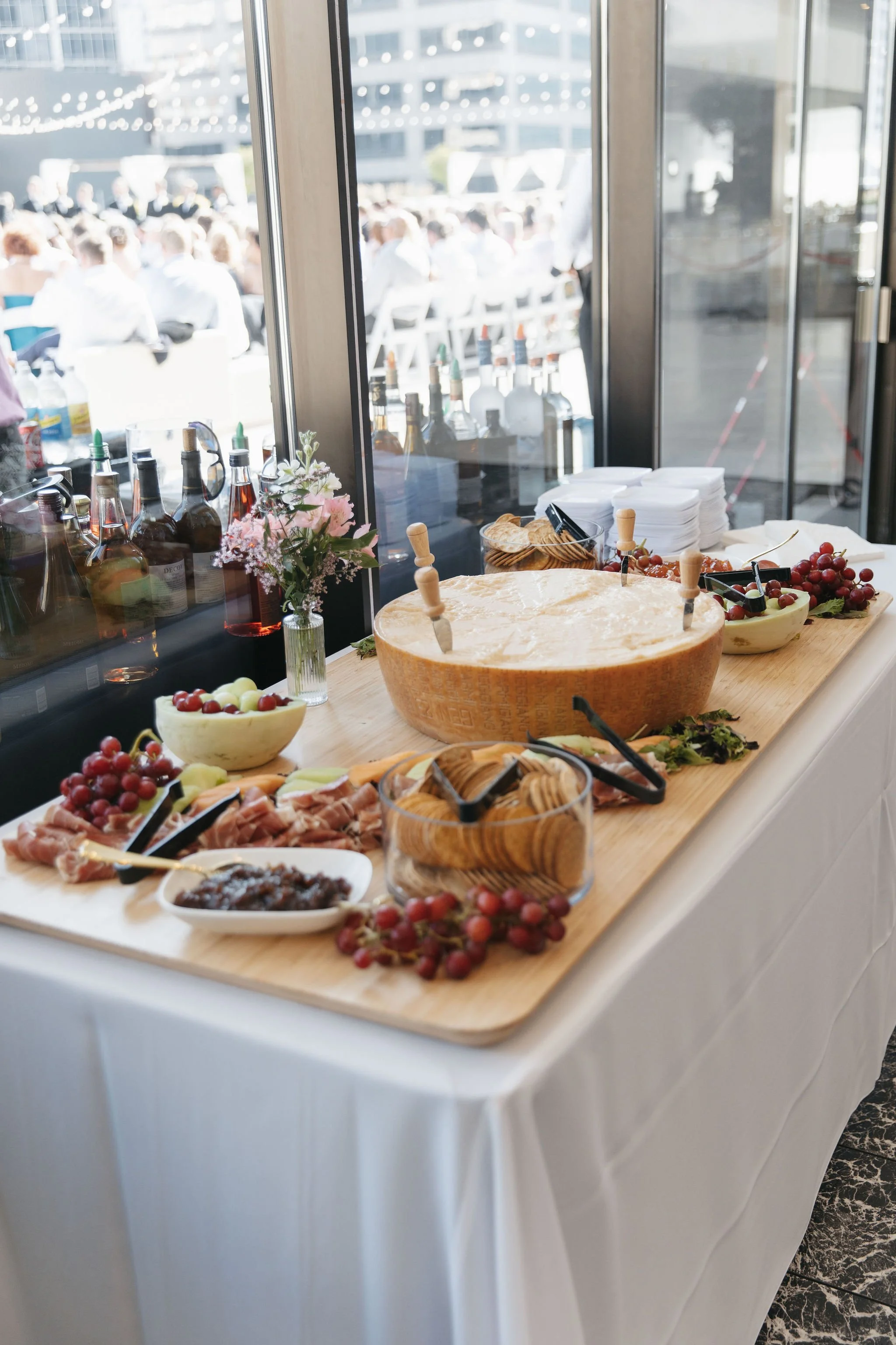A buffet table with various foods including grapes, sliced meats, cheese, and spreads, set against large window with an outdoor event in the background.