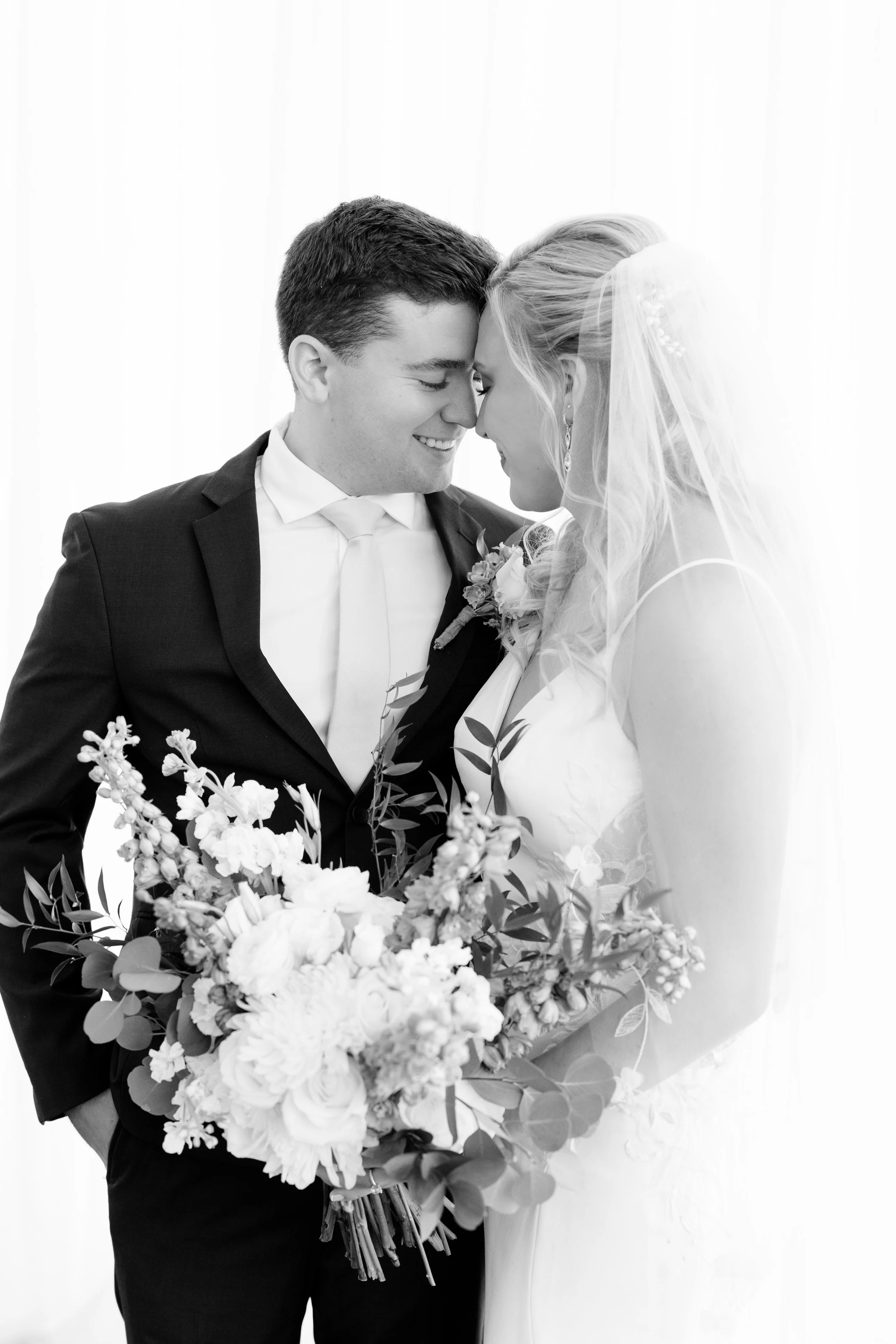 A black-and-white photograph of a newlywed couple, with their foreheads touching and eyes closed, holding a large bouquet of flowers, against a plain light background.
