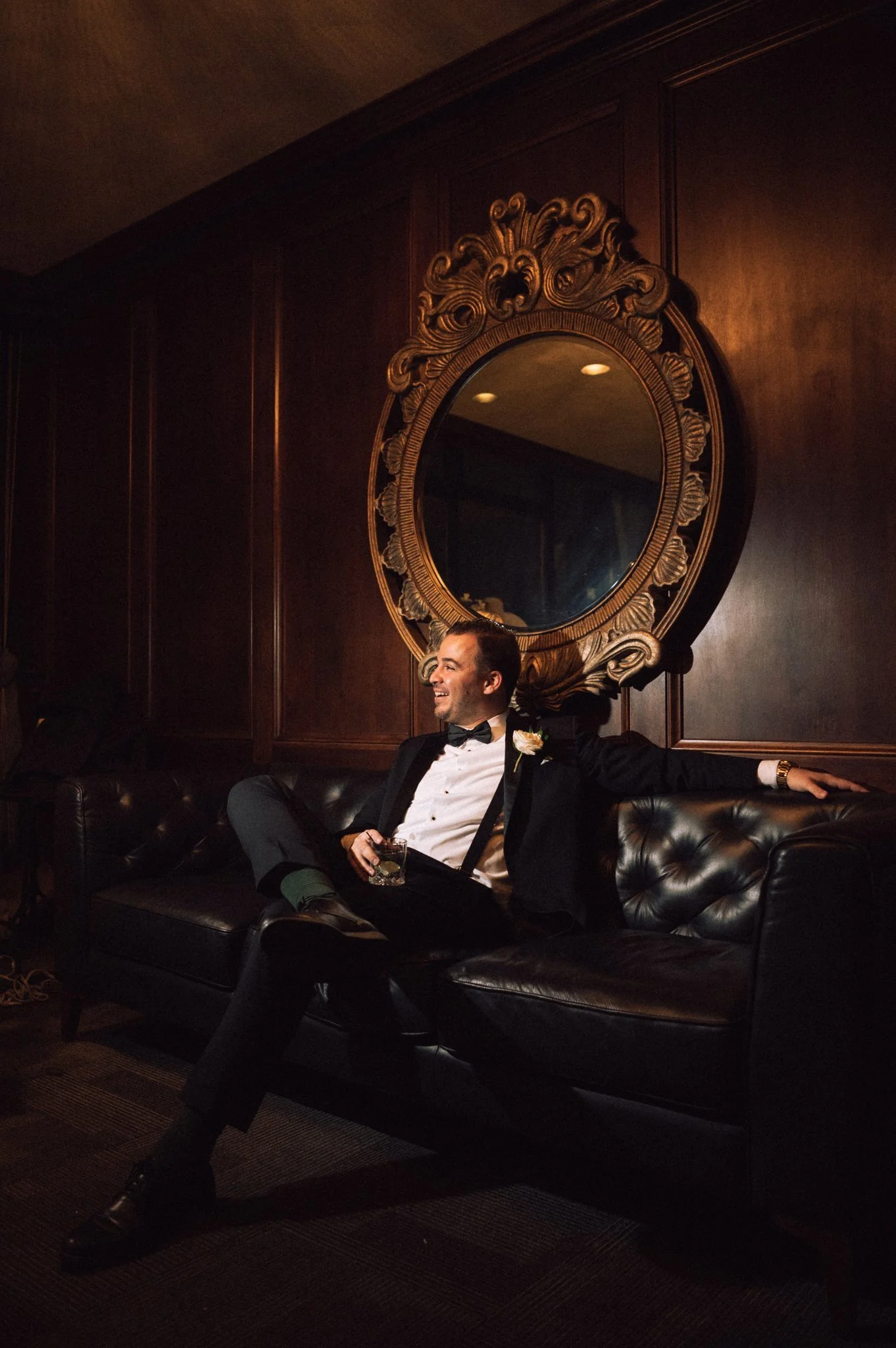 A man in a tuxedo sitting on a black leather couch in a dimly lit room. He is smiling, holding a drink, and leaning back with his arms stretched out. There is a large ornate mirror on the dark wood-paneled wall behind him.