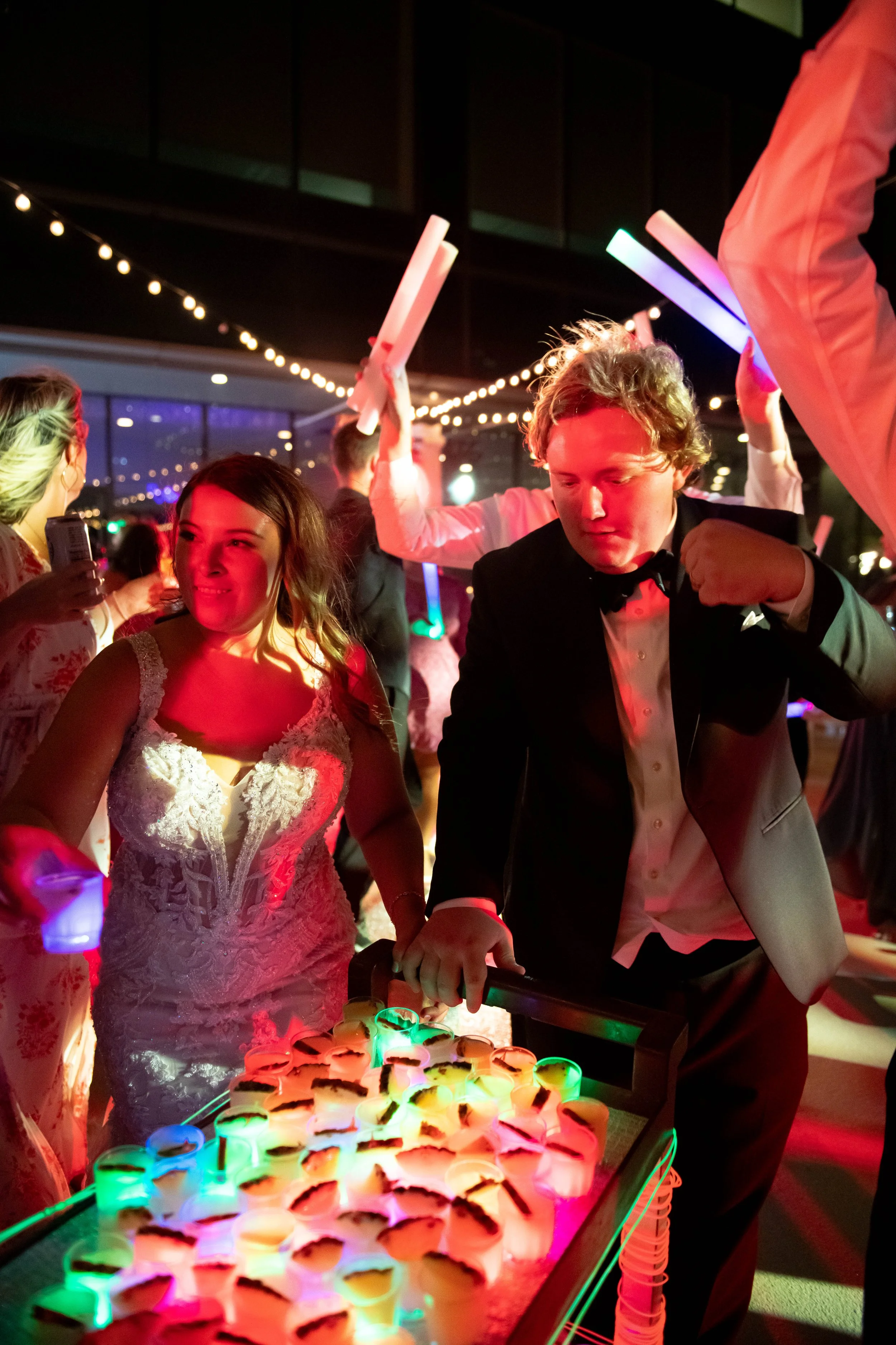 People playing a glow-in-the-dark beer pong game at a party, illuminated by colorful LED lights, with a woman in a white lace dress and a man in a tuxedo.
