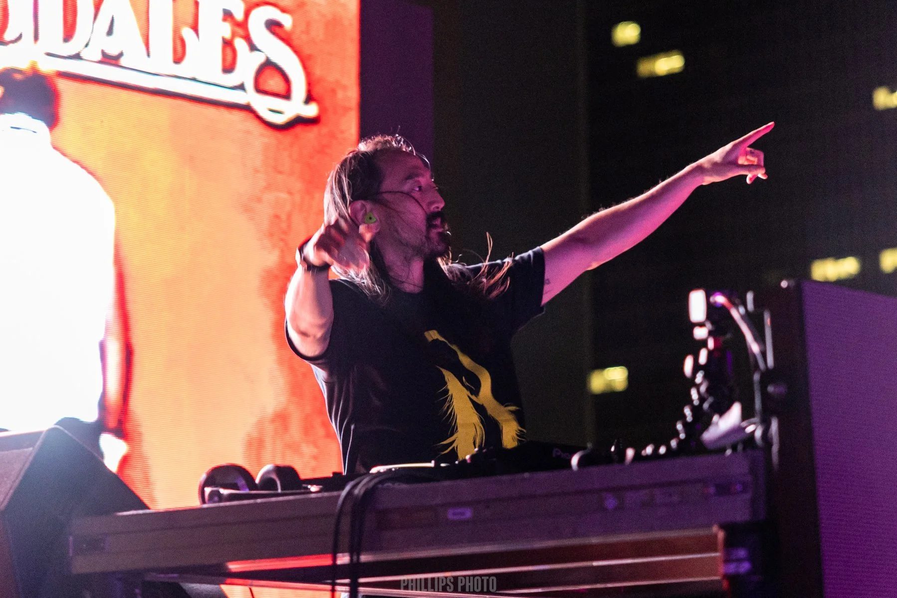A DJ with long hair, wearing a black T-shirt with a yellow graphic, pointing to the right while performing at a live event. Large electronic screens with orange and purple visuals are in the background.