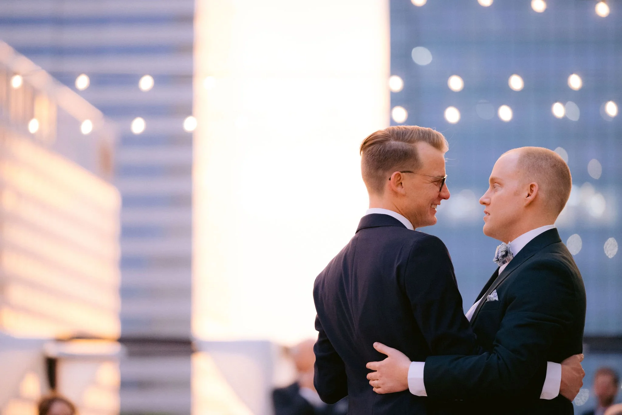 Two grooms embracing at sunset under string lights on the 11,000 sq ft terrace at JPS Events at Regions Tower. An inclusive, LGBTQ+-friendly luxury venue with 360 Indy skyline views & zero late-out fees.