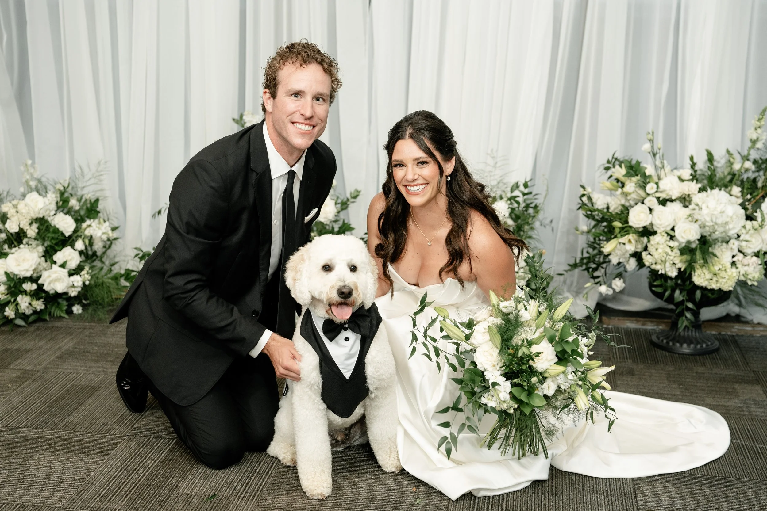 Dog-friendly wedding couple with their pet in a tuxedo posing before a white rose floral wall at JPS Events at Regions Tower. Highlights in-house wholesale decor & full-service planning in downtown Indy.