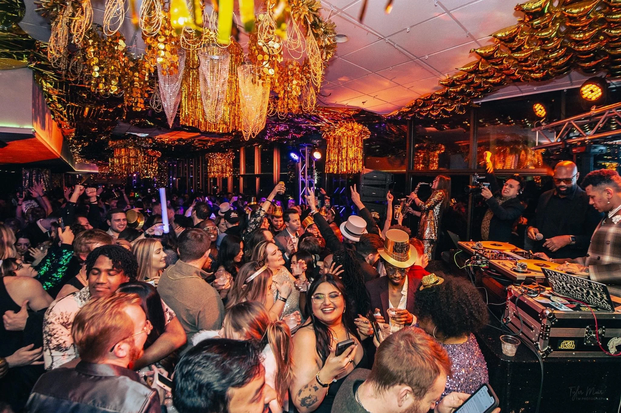 Crowd of people dancing and enjoying music at a lively party or celebration with gold decorations hanging from the ceiling and colorful lighting.