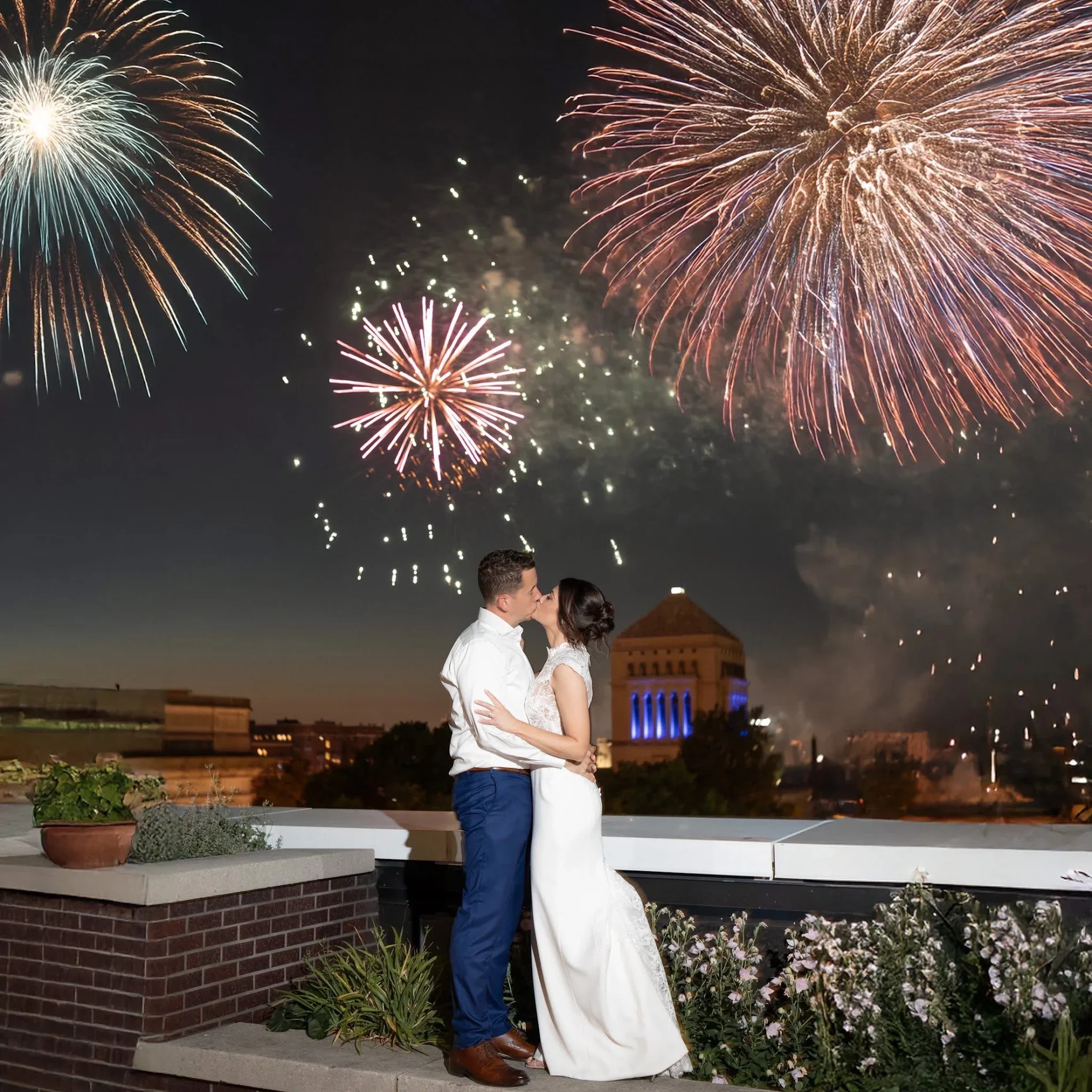 A couple in formal wedding attire kissing under fireworks on the private exclusive 5th-floor rooftop terrace at JPS Events at Regions Tower in Downtown Indianapolis, with city skyline buildings and lush plants illuminated in the background.