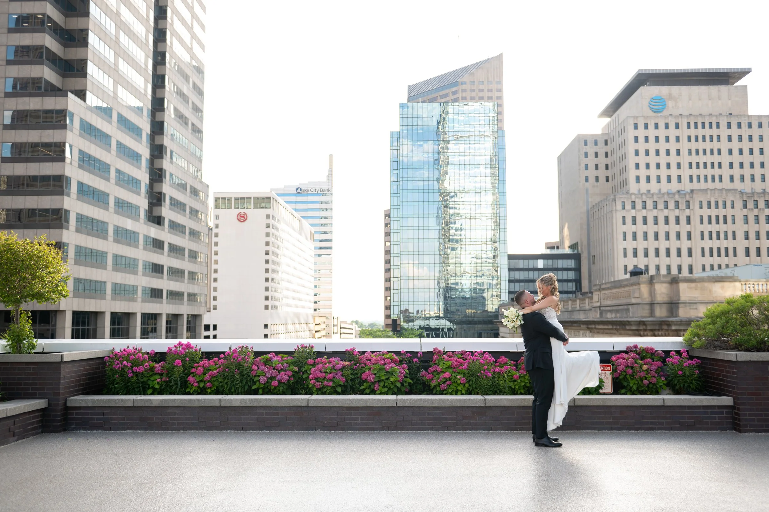 A newlywed couple sharing a kiss on a rooftop with a city skyline and tall buildings in the background, flowers in the foreground, and a bright sky.