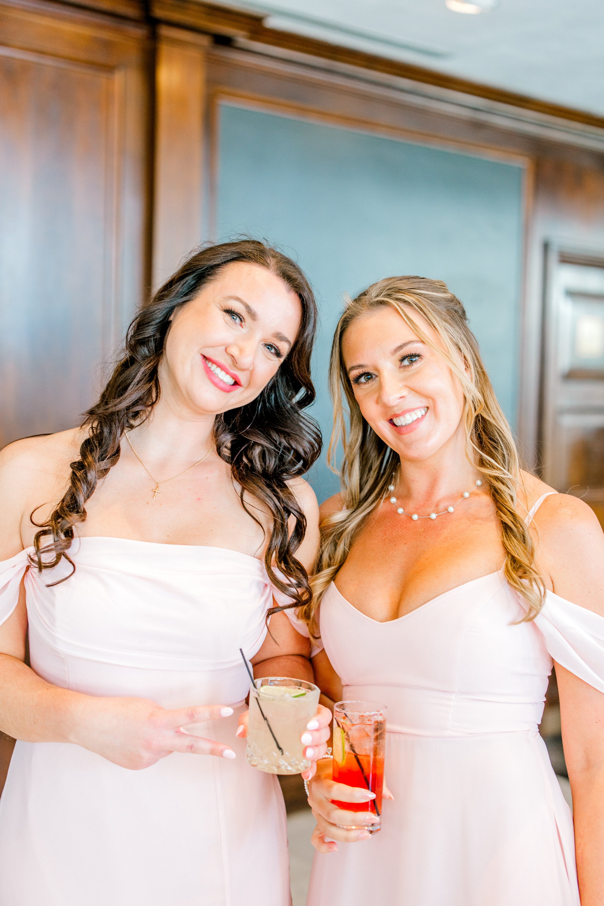 Two women wearing matching pink dresses, smiling, holding cocktails, standing indoors with wooden walls and a blue wall in the background.