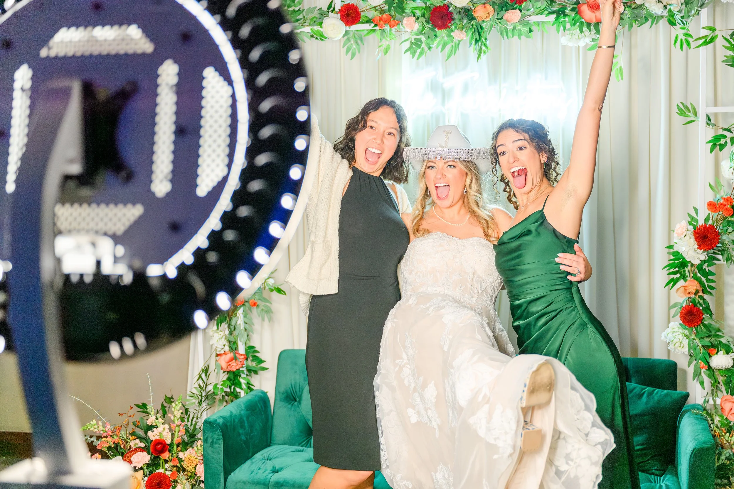 Three women celebrating at a wedding, one in a white wedding dress with a veil, holding up her arm, another in a green dress with her tongue out and a third in black with a wide smile, all standing in front of a floral backdrop.