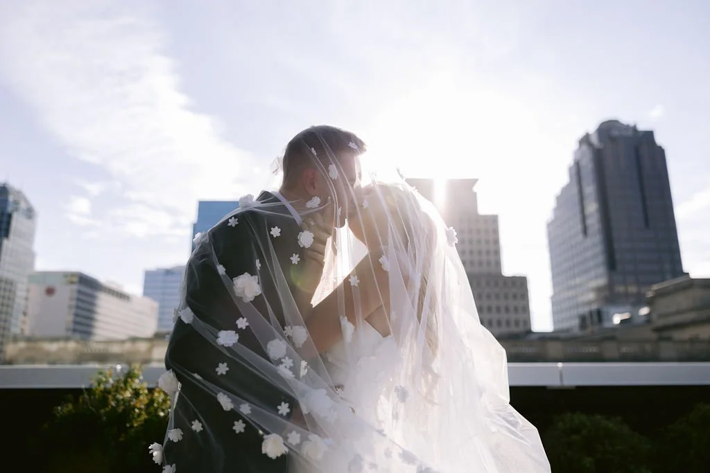 A couple in wedding attire sharing a kiss under a veil with floating flowers, with a city skyline and sunlight in the background.