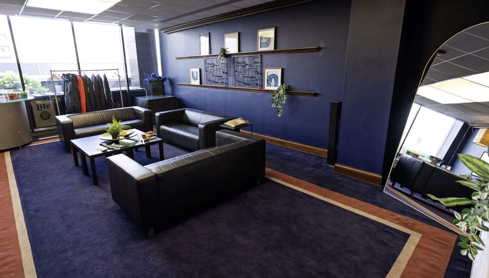 A modern lounge area with three black leather sofas, a black coffee table with a potted plant, and a dark blue wall decorated with framed pictures and hanging plants. In the background, there are clothing racks, a table with blue flowers, and a mini fridge, with sunlight coming in through large windows.
