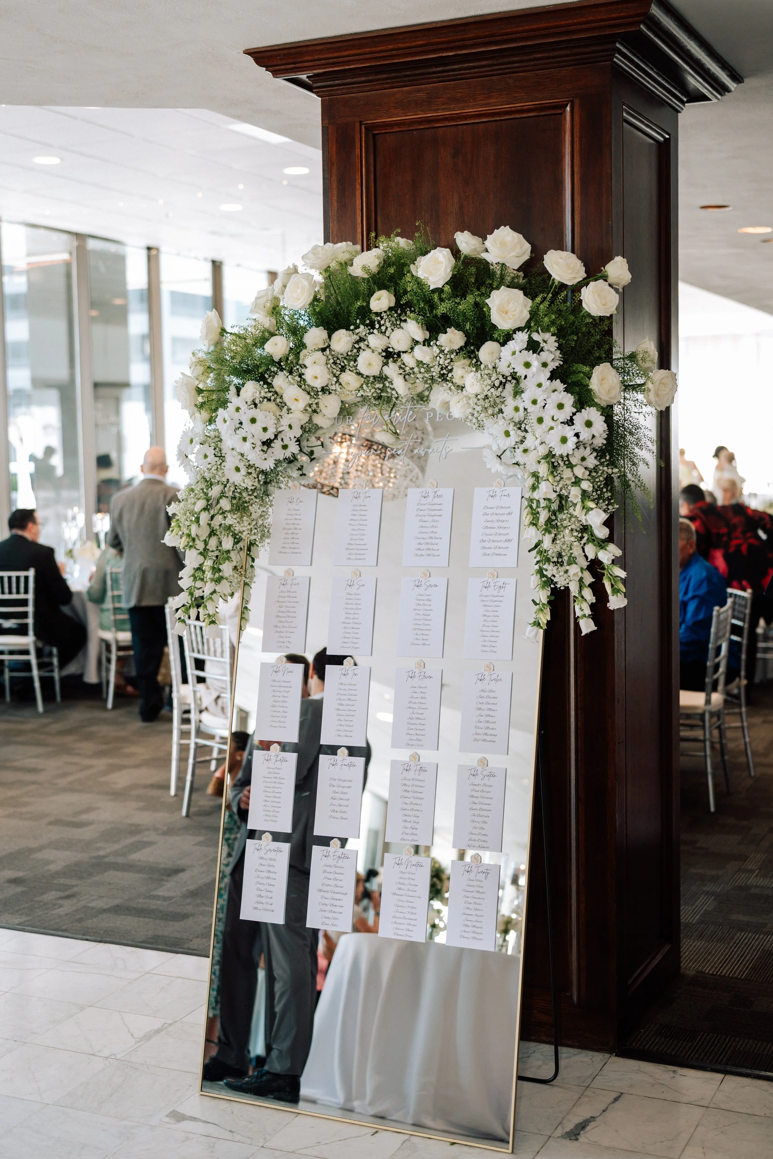 A large wedding seating chart with a floral arrangement on top, featuring white roses and daisies. The chart displays multiple white sheets with table assignments, positioned on an easel. The setting appears to be a reception area with people seated 