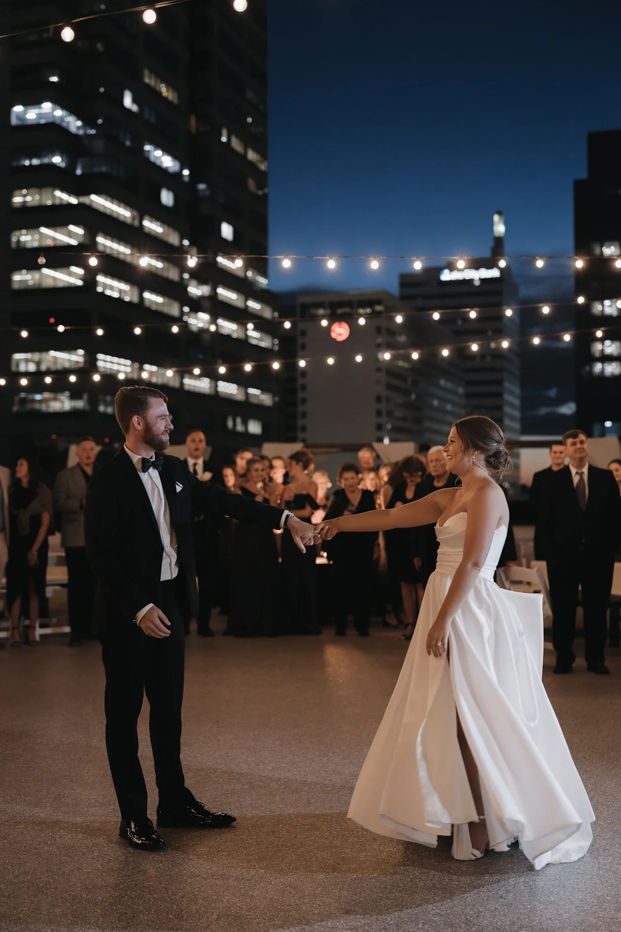 A bride and groom dancing at their night wedding reception on the 11,000 sq ft private rooftop terrace at JPS Events at Regions Tower in Downtown Indianapolis, surrounded by guests, string lights, and illuminated skyline views.