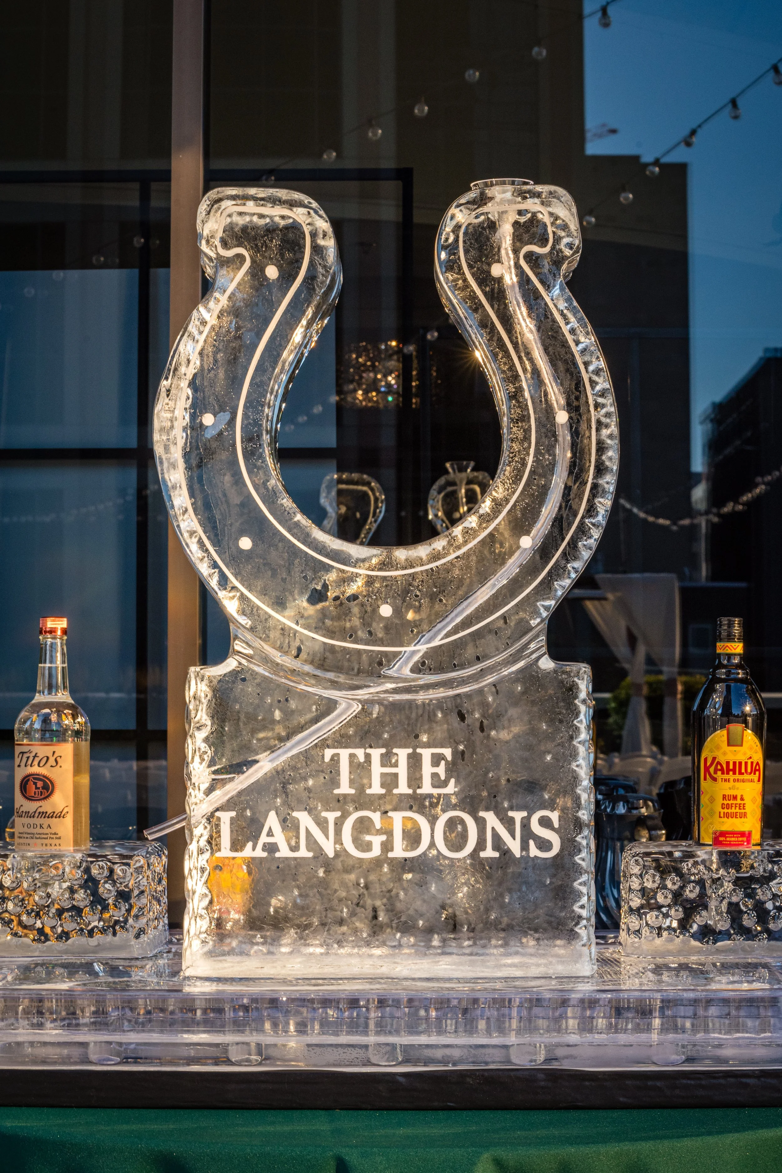 Ice sculpture with the words "The Lángdons" engraved, illuminated, with bottles of liquor on either side and decorative elements in the background.
