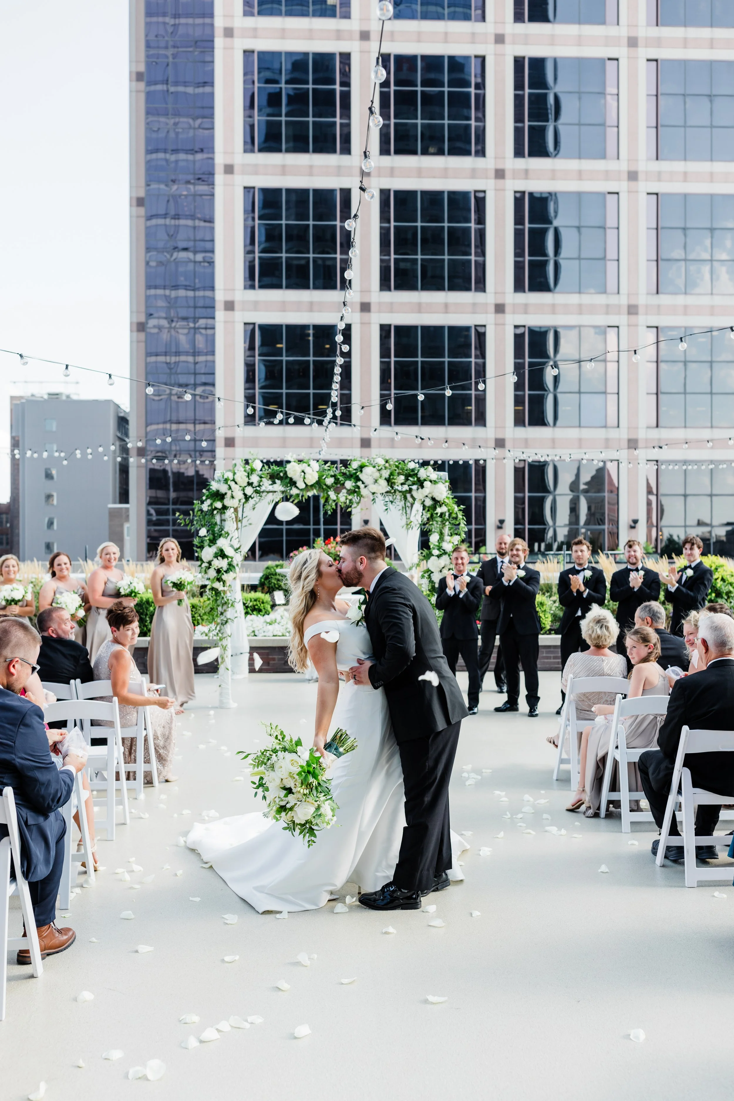 A couple in wedding attire kissing during their outdoor ceremony on the private 11,000 sq ft rooftop terrace at JPS Events at Regions Tower in Downtown Indianapolis, with guests on white chairs and city skyline views.