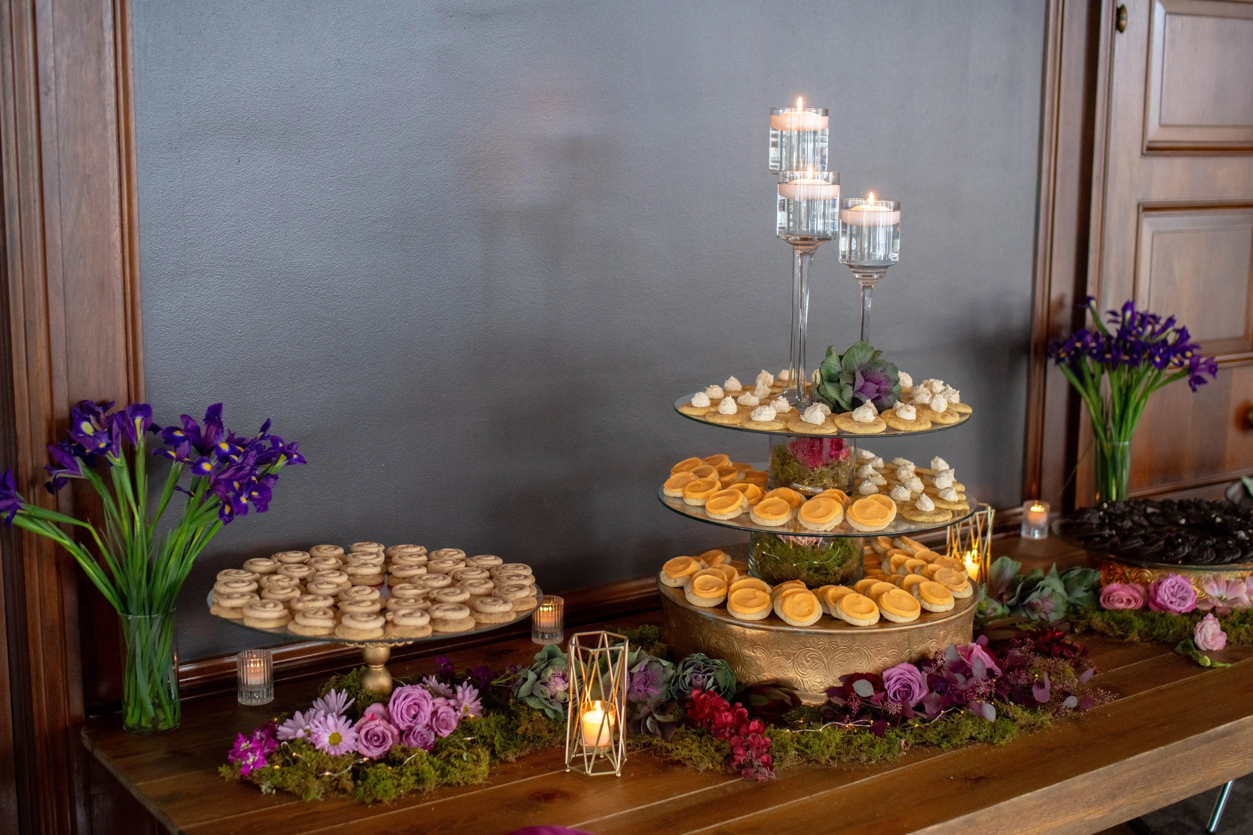 A dessert table with a multi-tiered display of cupcakes and cookies, surrounded by purple flowers, candles, and greenery, against a gray wall and wooden cabinetry.