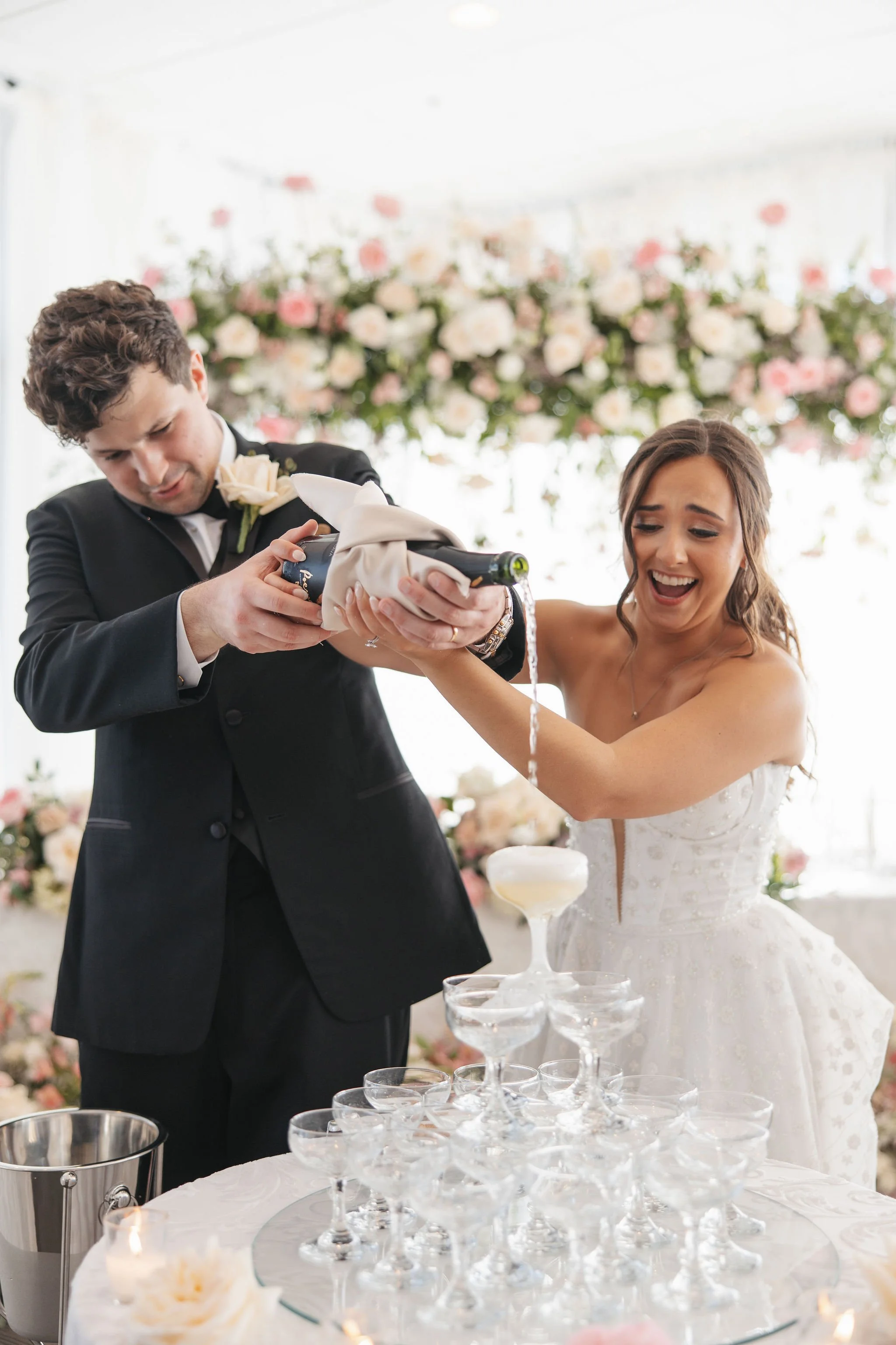 A bride and groom at a wedding, pouring champagne into a glass tower, with a floral backdrop.