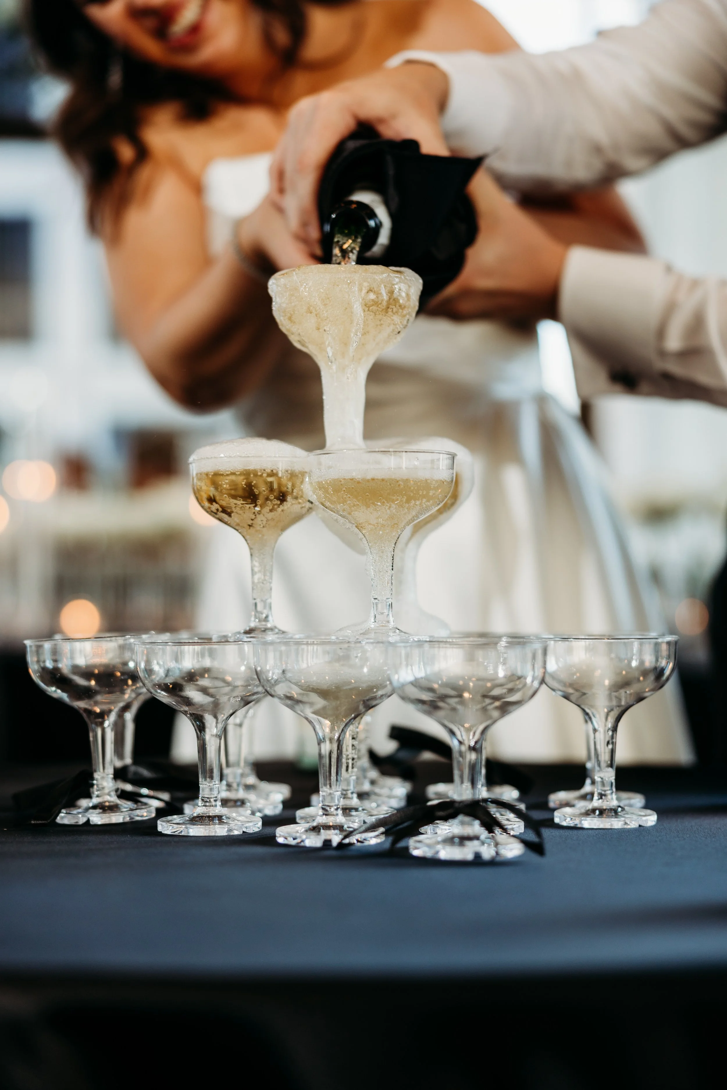 A woman in a white dress is pouring champagne from a bottle into a pyramid of coupe glasses at a celebration or event.