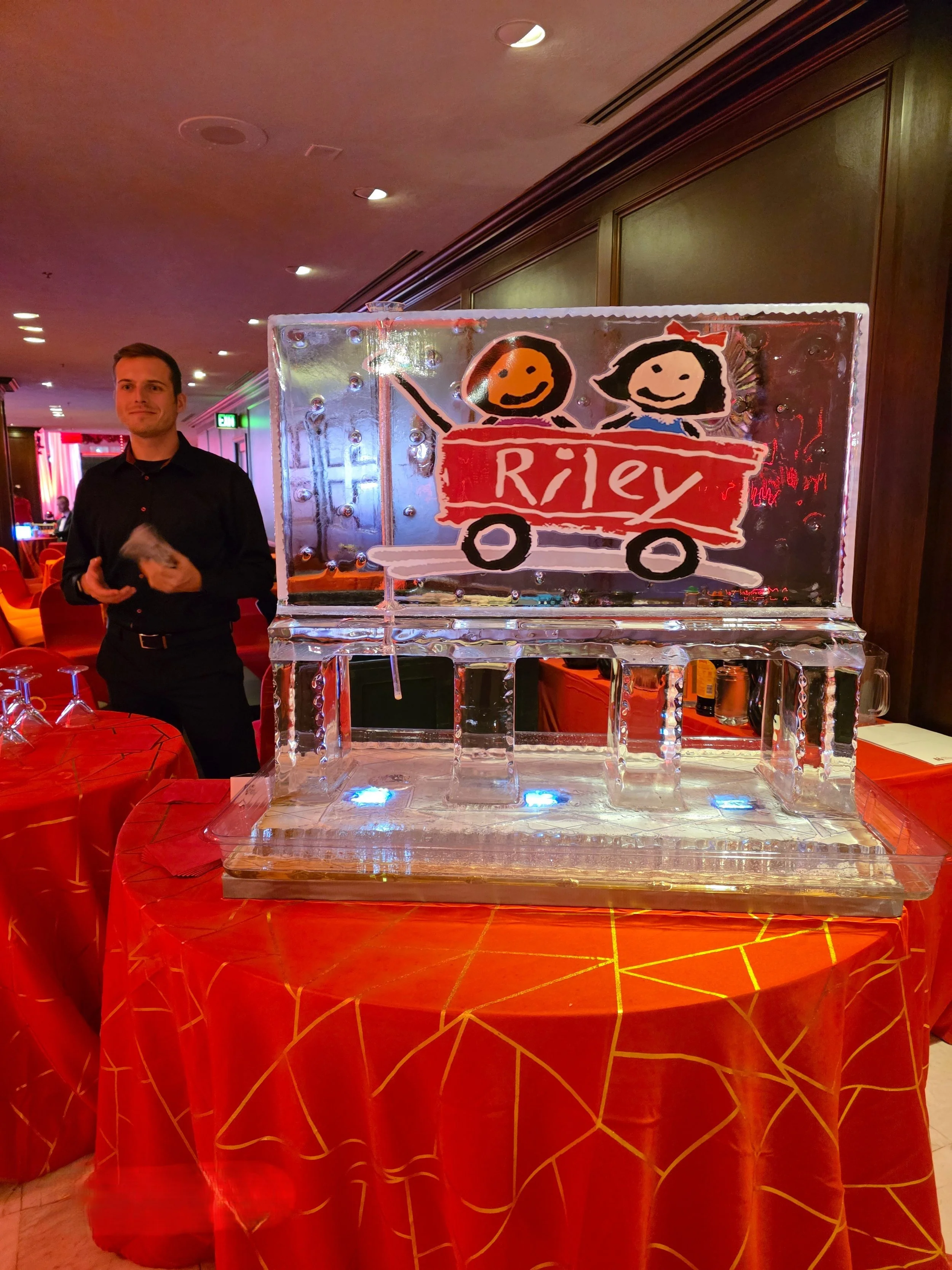 Ice sculpture of two children on a school bus with the word 'Riley' written on it, displayed at an indoor event with red tablecloths and a man standing nearby.