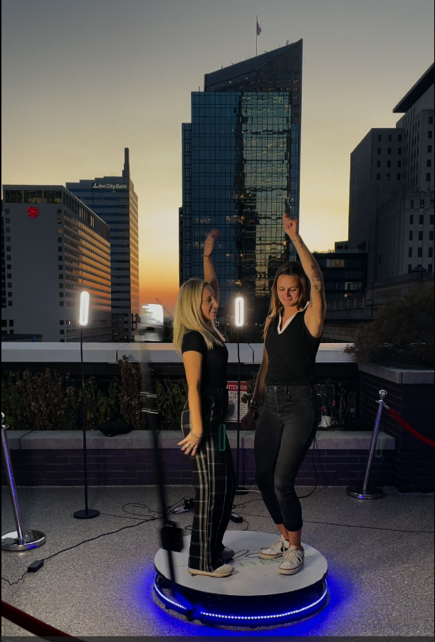 Two women dancing on a small illuminated circular stage outdoors in a city skyline at sunset, with tall buildings and modern architecture in the background.