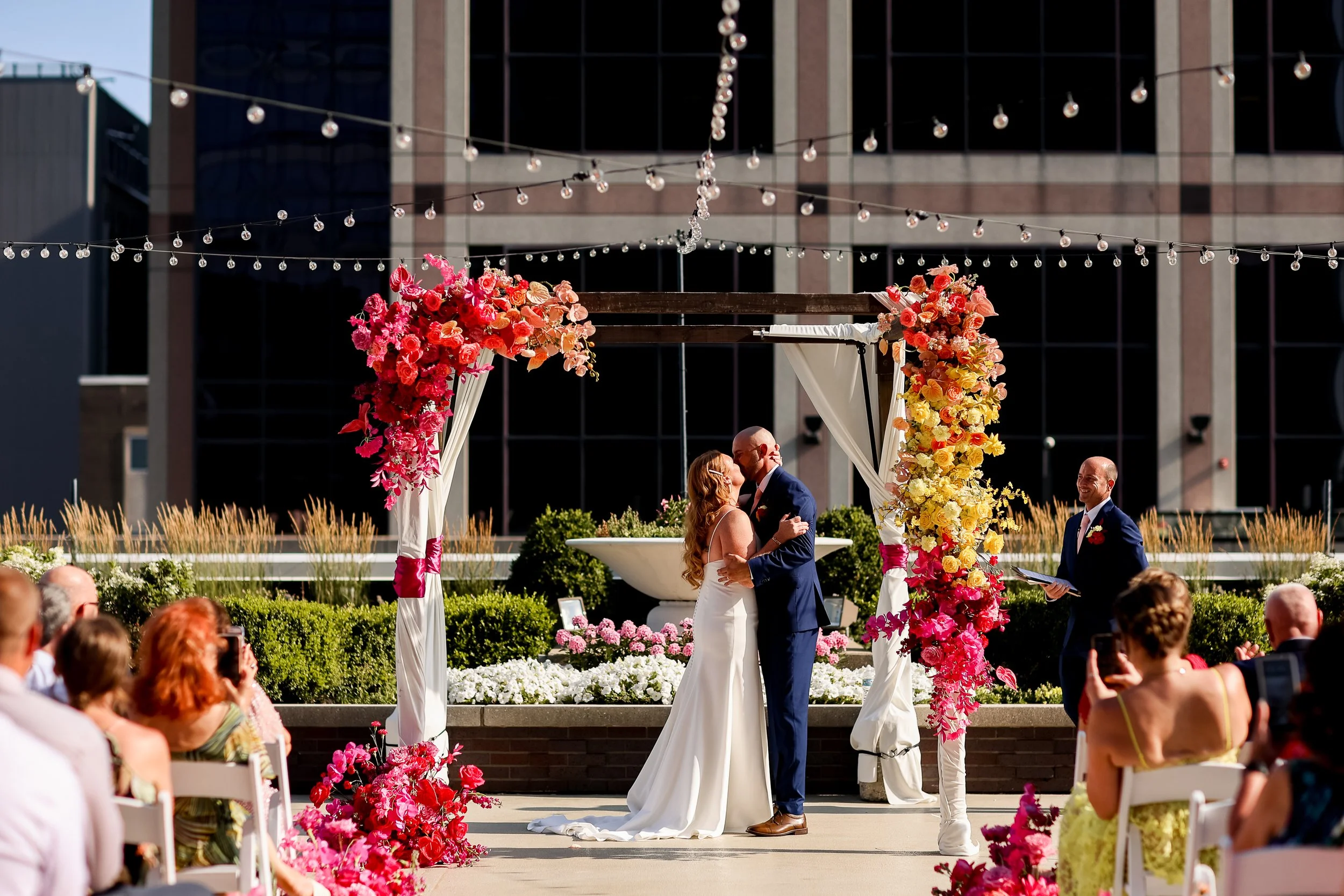 A couple getting married under a floral arch at an outdoor wedding ceremony, with guests watching and taking photos.