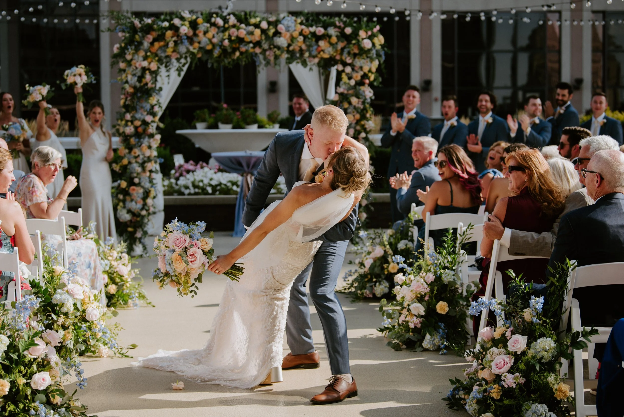 A newlywed couple shares a kiss at their outdoor ceremony on the private exclusive rooftop terrace at JPS Events at Regions Tower in Downtown Indianapolis, with guests cheering and custom in-house floral decorations lining the aisle.